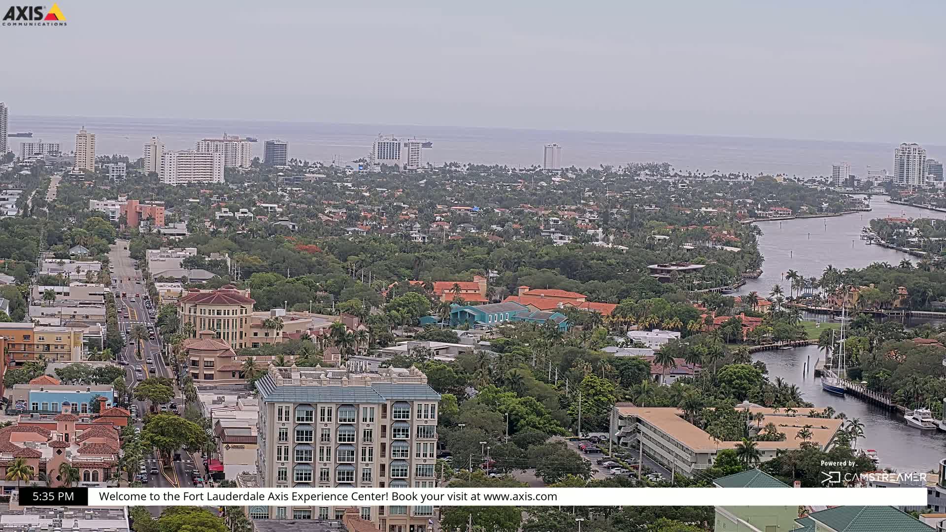 An aerial view of a coastal city on an overcast day, showing a network of waterways, buildings, and lush green landscaping extending to a calm ocean horizon.