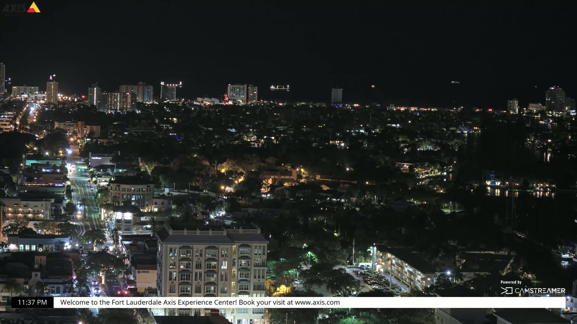 A nighttime aerial view of a city with brightly lit buildings and streets, extending to a dark coastline and water beyond.