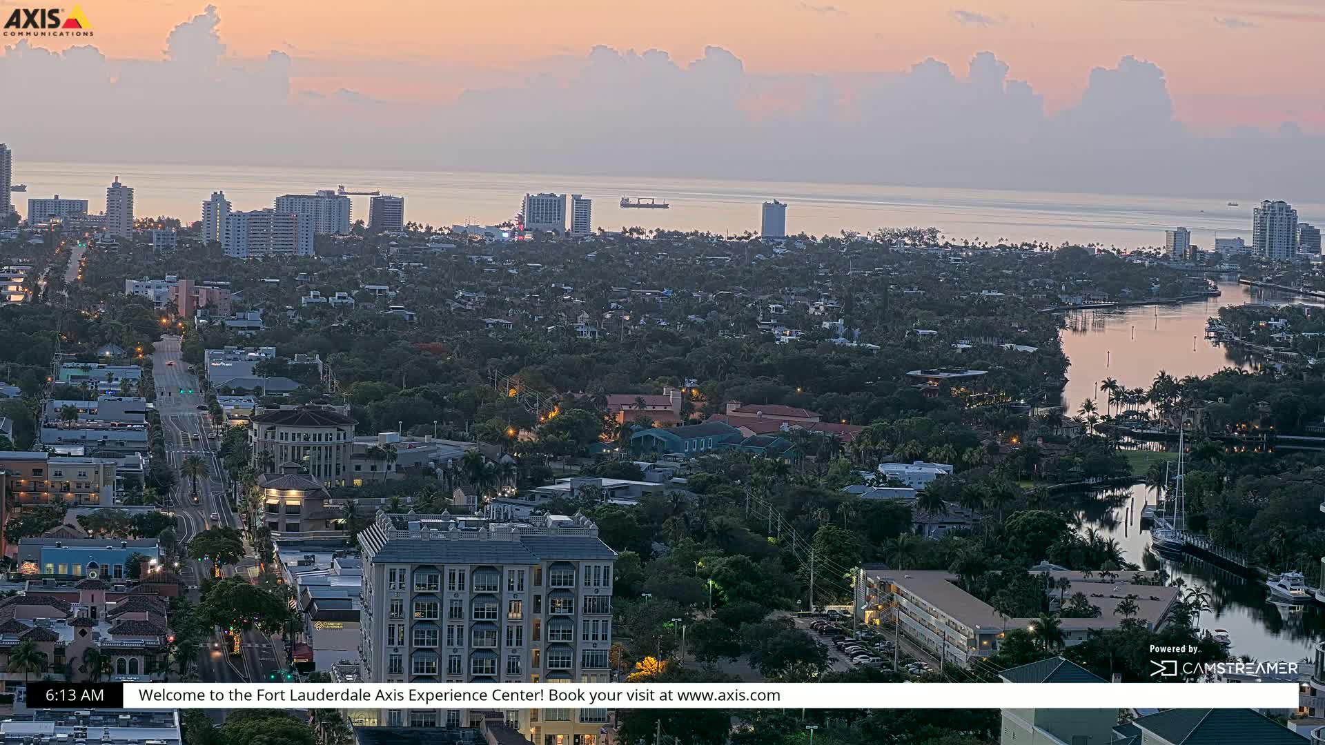 A coastal city at sunrise, with calm waters reflecting the pale pink and orange sky, is viewed from a high vantage point overlooking tree-lined streets, buildings, and a waterway.