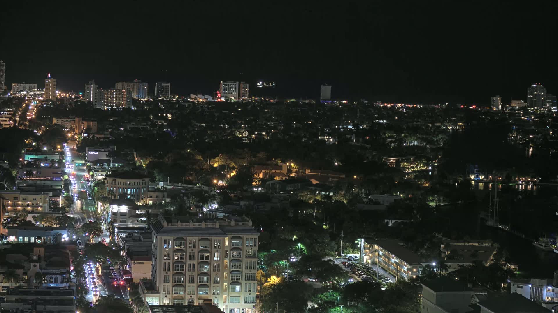 A nighttime aerial view of a city with a main road illuminated by traffic, surrounded by residential buildings and lush greenery, extending to a dark waterfront with distant city lights and a dark sky.