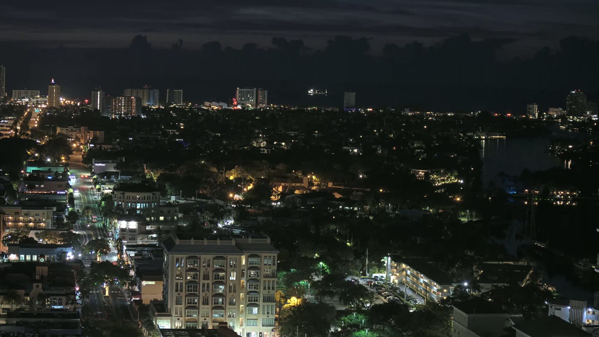 A nighttime aerial view of a city with a dark sky showing a cityscape illuminated by streetlights and building lights, including a body of water in the distance.