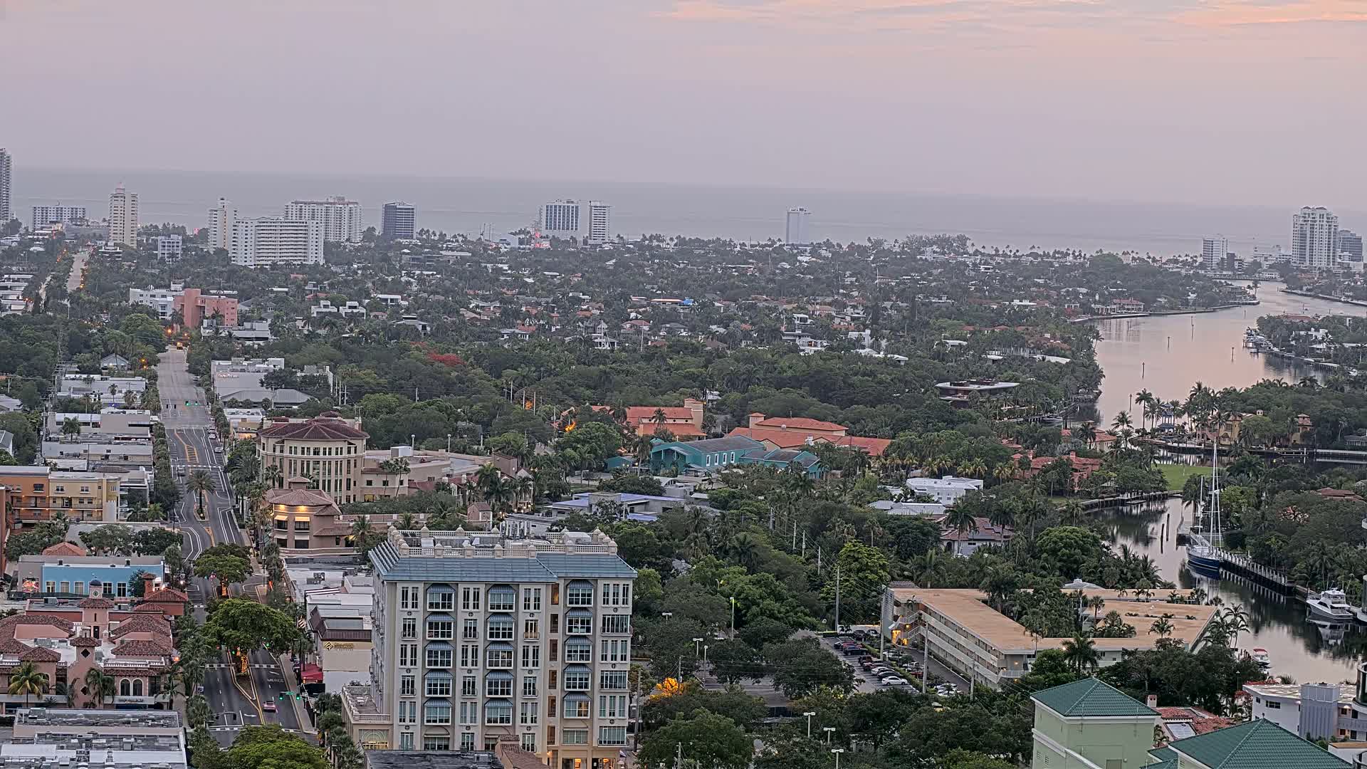 A coastal city at dawn, with calm waters reflecting the pale sky, shows a mix of residential buildings and high-rises along a waterway.