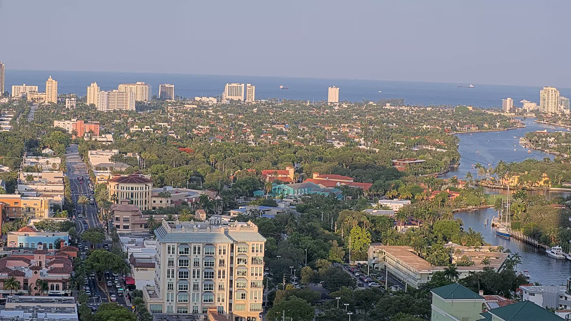 An aerial view shows a coastal city with a waterway winding through a dense, lush landscape under a clear, sunny sky.