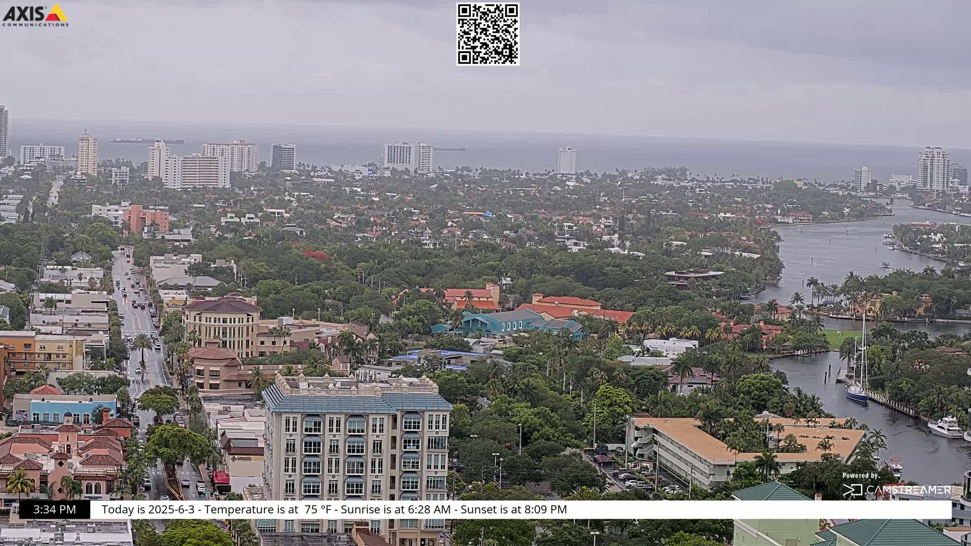 A coastal city on an overcast day is seen from an elevated viewpoint, showcasing a waterway winding through a dense residential area with several high-rise buildings visible in the distance.