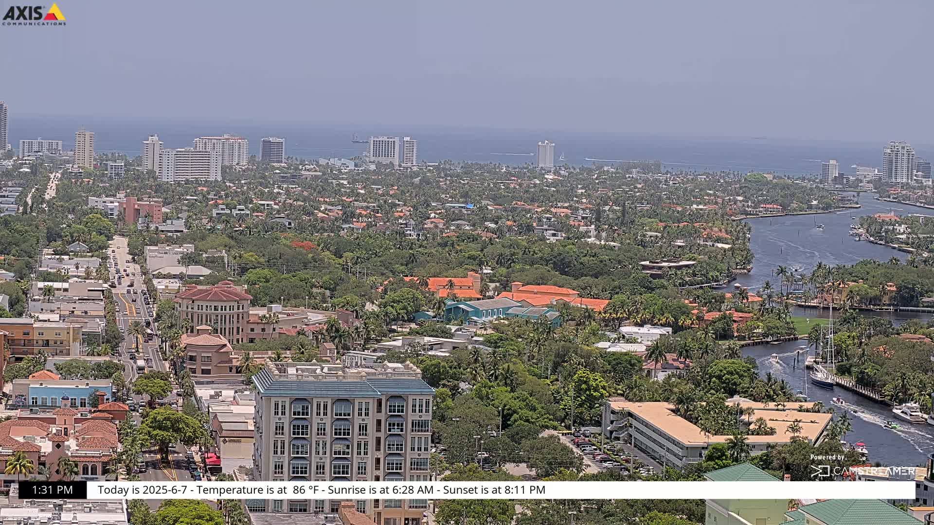 A hazy, sunny day reveals a coastal city scene with numerous buildings, waterways, and lush greenery, extending to a distant ocean.