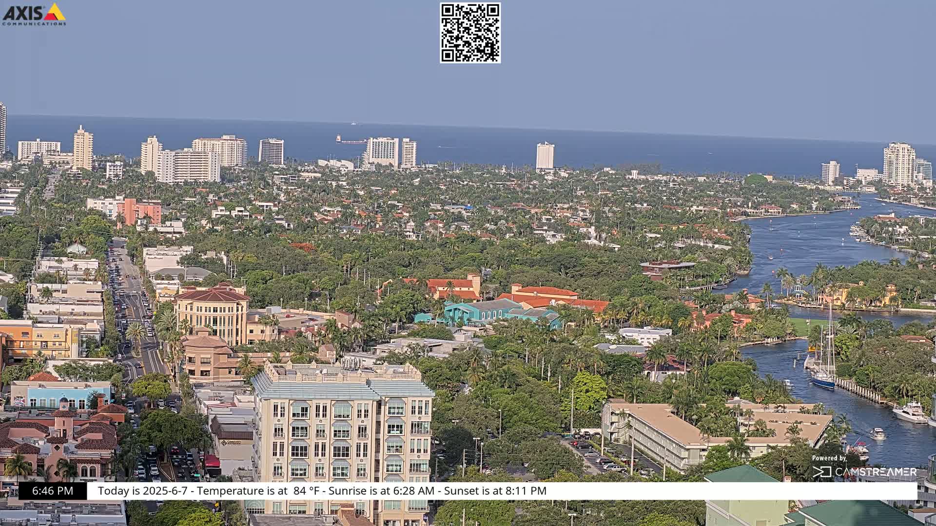 A high-angle, long shot of a coastal city on a sunny day shows a waterway winding through a neighborhood of houses and low-rise buildings towards the ocean in the distance.