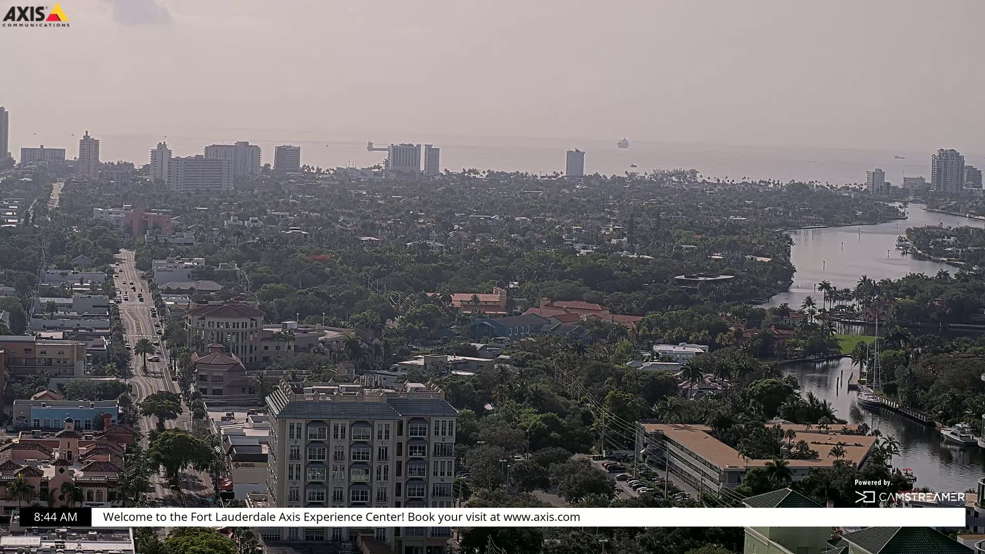 A hazy, sunny day reveals a sprawling cityscape with a waterway winding through a dense residential area, leading to a distant ocean with a few visible ships.