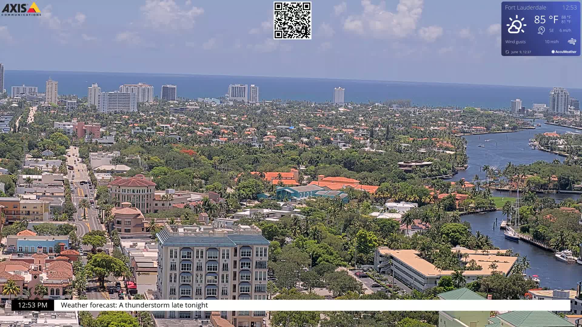 A high-angle view of a coastal city on a partly sunny day, with a body of water and numerous buildings visible, shows a forecast of thunderstorms later that evening.