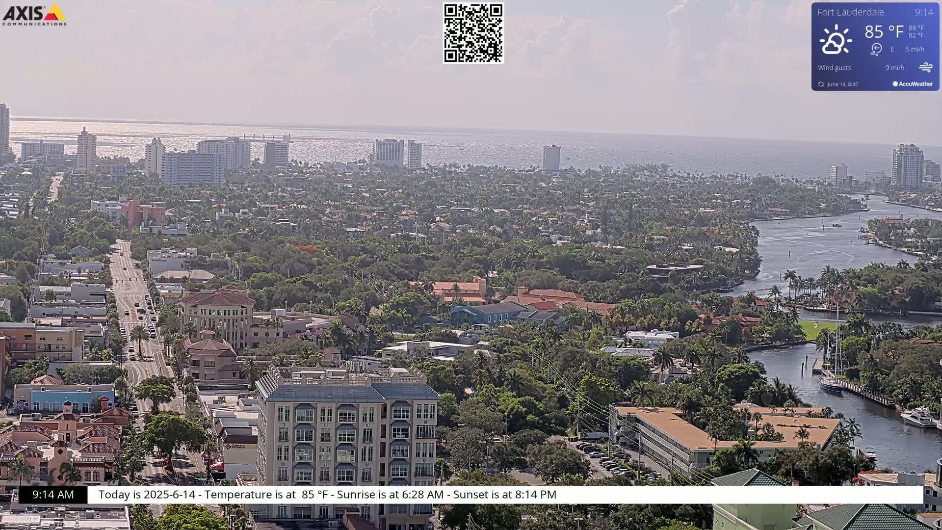 An aerial view of a coastal city on a partly sunny day, showing buildings, roads, waterways, and lush vegetation.