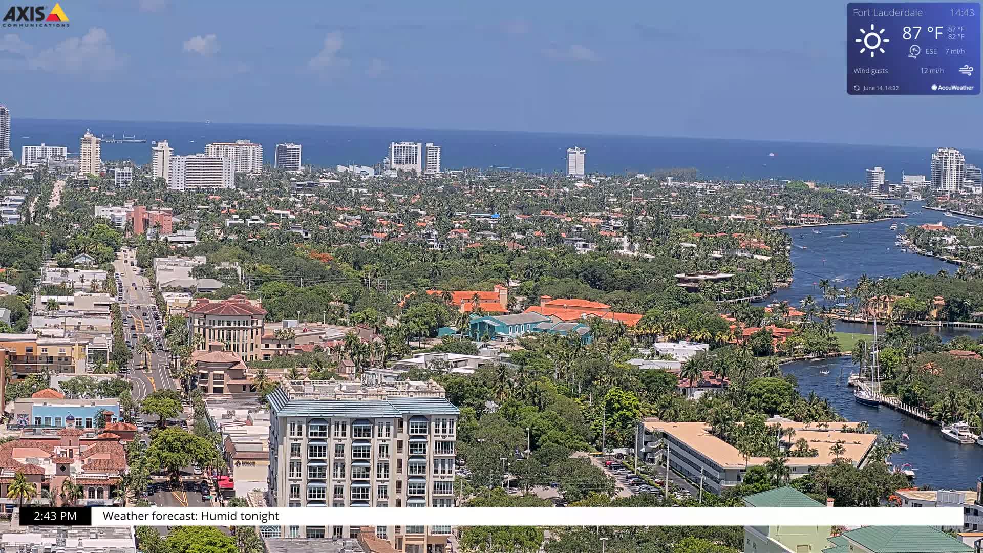 An aerial view of a coastal city on a partly sunny day, showing buildings, roads, waterways, and lush vegetation.