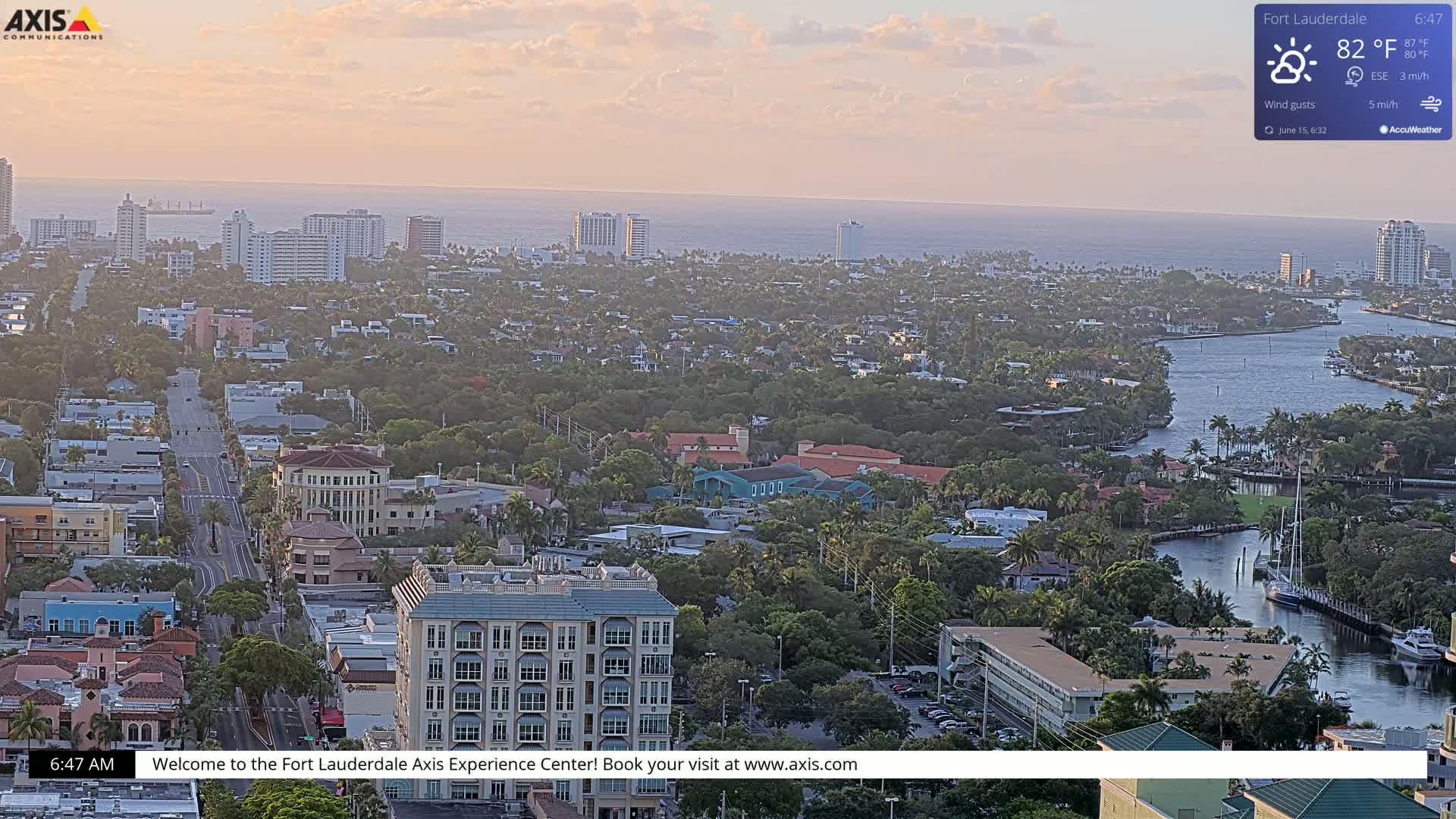 An aerial view of a coastal city at sunrise, showing a waterway winding through a residential area towards the ocean under partly cloudy skies.