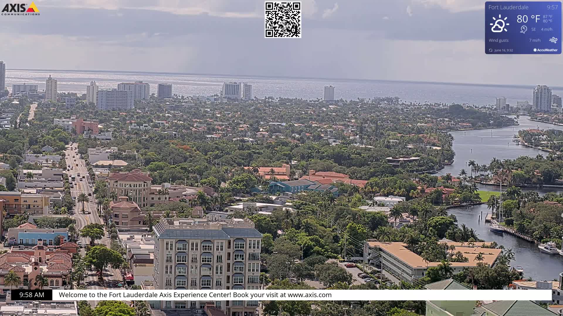 An aerial view of a coastal city on a partly cloudy day shows a waterway winding through a lush, green landscape of buildings and trees, extending to the ocean in the background.
