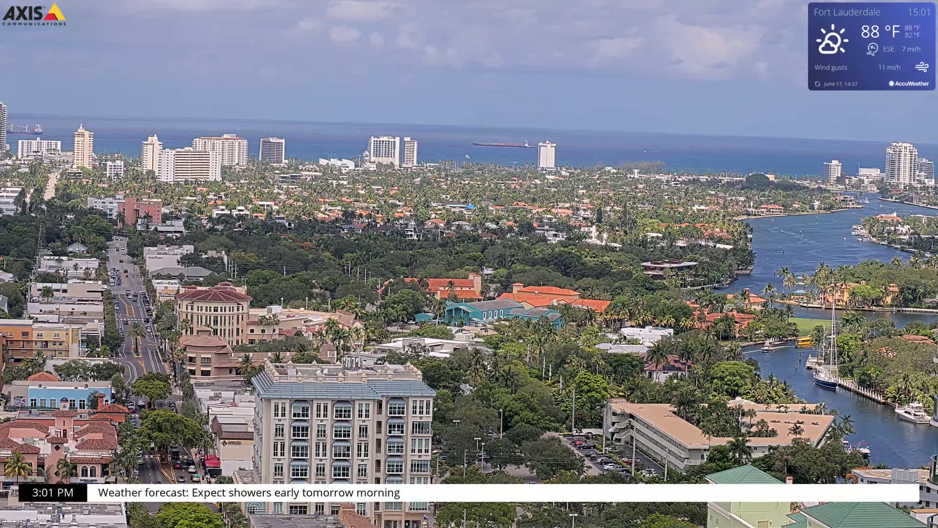 An aerial view of a coastal city with a waterway, featuring many buildings and lush greenery under partly cloudy skies.