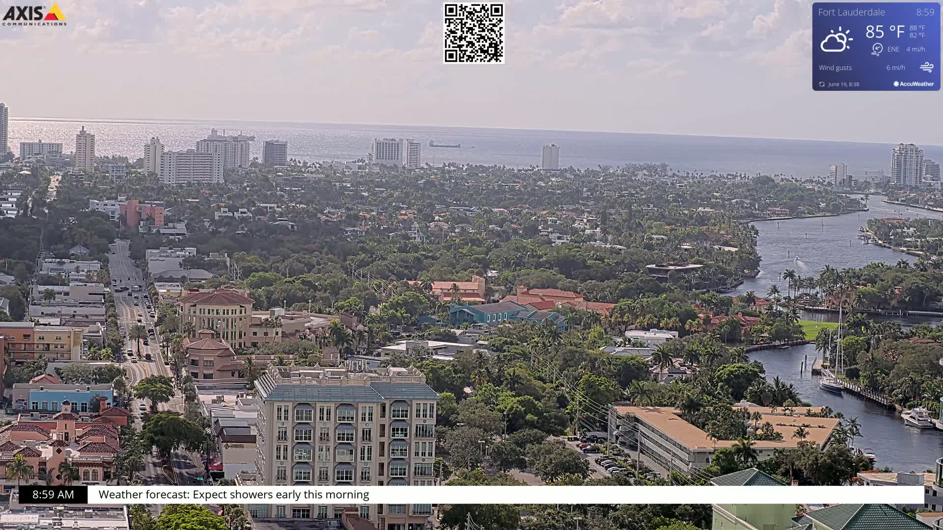 An aerial view shows a coastal city with a waterway winding through residential areas and high-rises, under partly sunny skies.