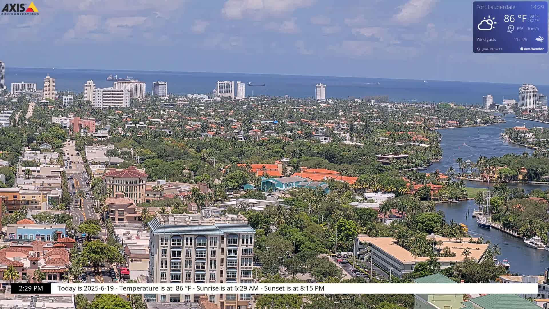 A sunny, coastal city with many buildings and waterways is viewed from above, with a partly cloudy sky.