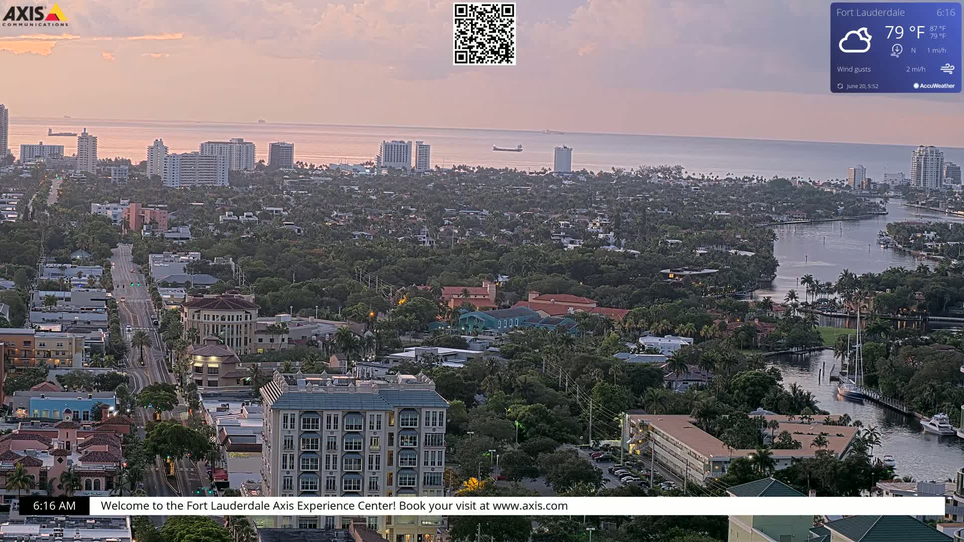 A high-angle, long shot of a city with calm waters and a partly cloudy sky at dawn shows buildings, roads, waterways, and lush vegetation.