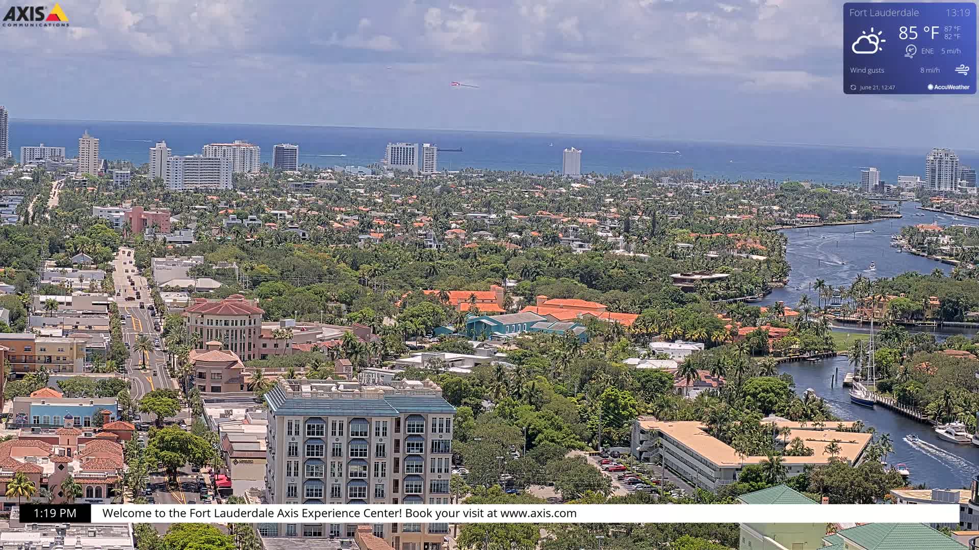 An aerial view of a coastal city on a partly sunny day shows a waterway winding through lush, green residential areas, with high-rise buildings and the ocean in the background.