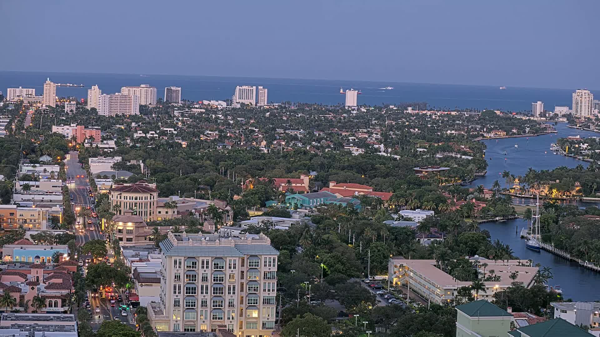 An aerial view at dusk shows a coastal city with buildings, streets, waterways, and lush vegetation, under a clear sky.