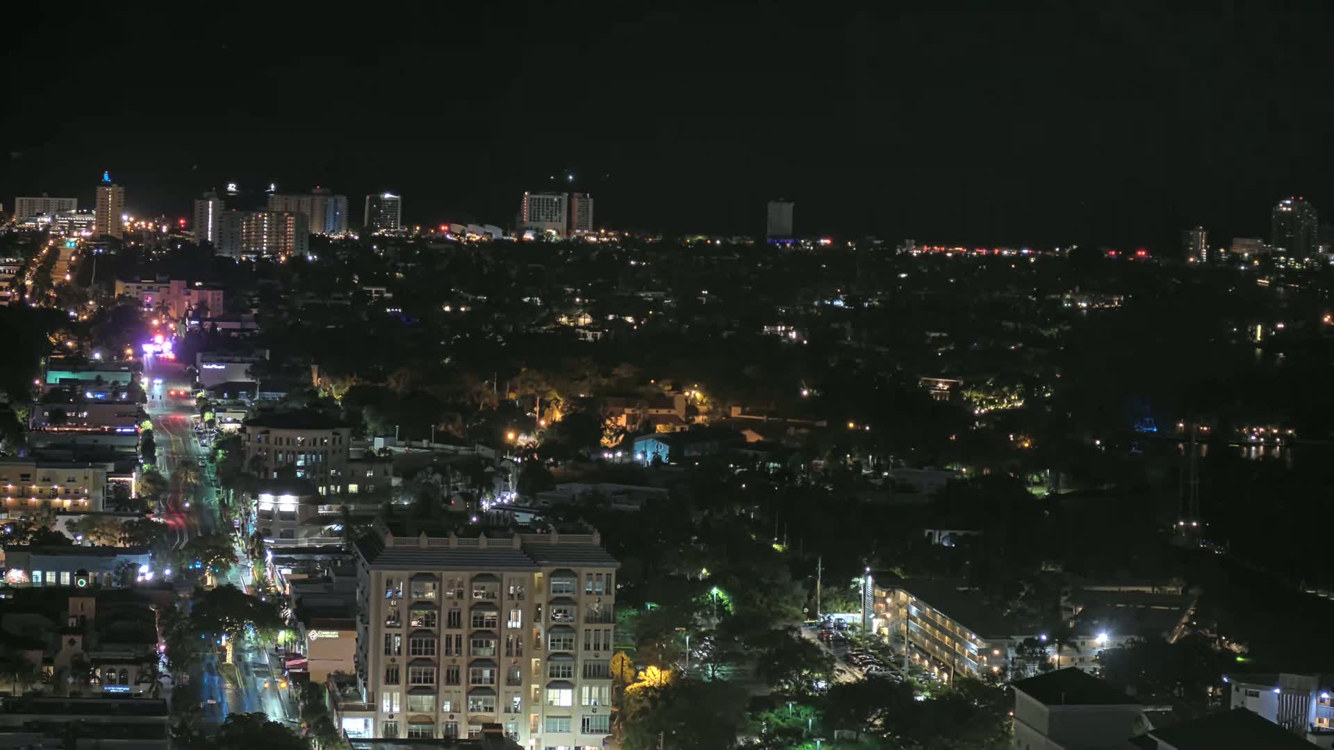 A nighttime aerial view shows a city with brightly lit streets and buildings, extending to a darker skyline of taller structures under a clear, dark sky.