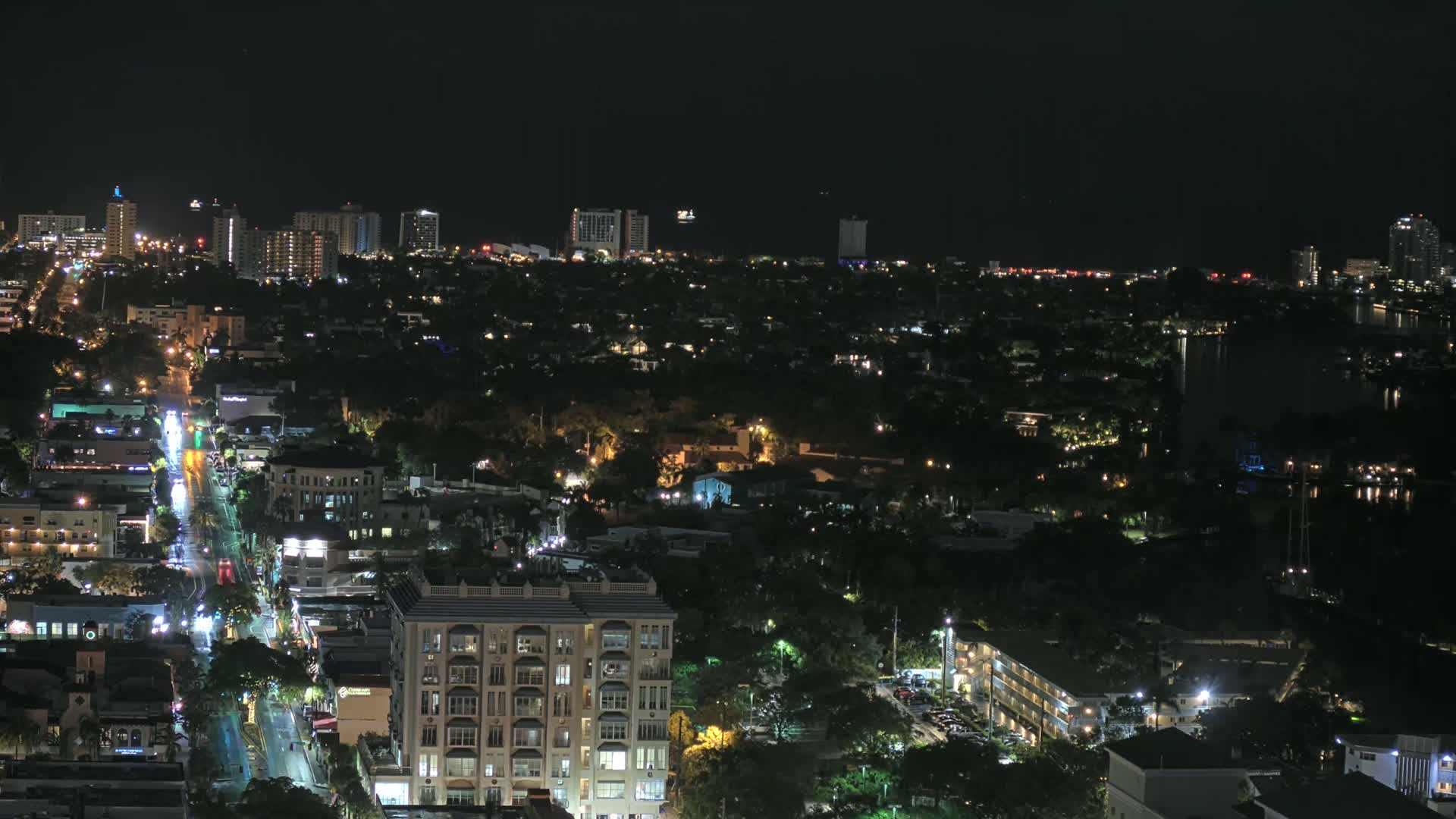 A nighttime aerial view shows a city's illuminated streets and buildings, with a darker waterfront area and a distant skyline visible under a clear, dark sky.