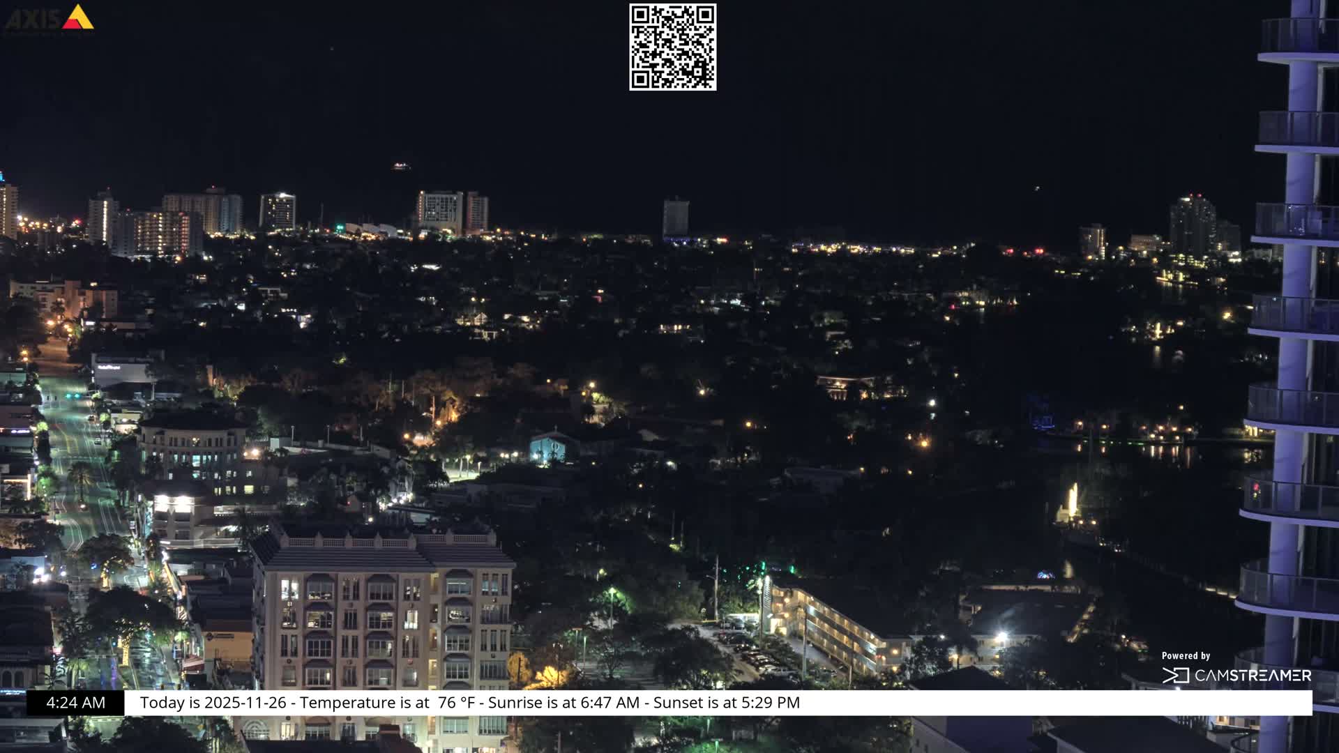 A clear night sky hangs over a brightly lit city featuring numerous buildings, streets, and a body of water reflecting lights, all viewed from an elevated perspective with a modern high-rise building framing the right.