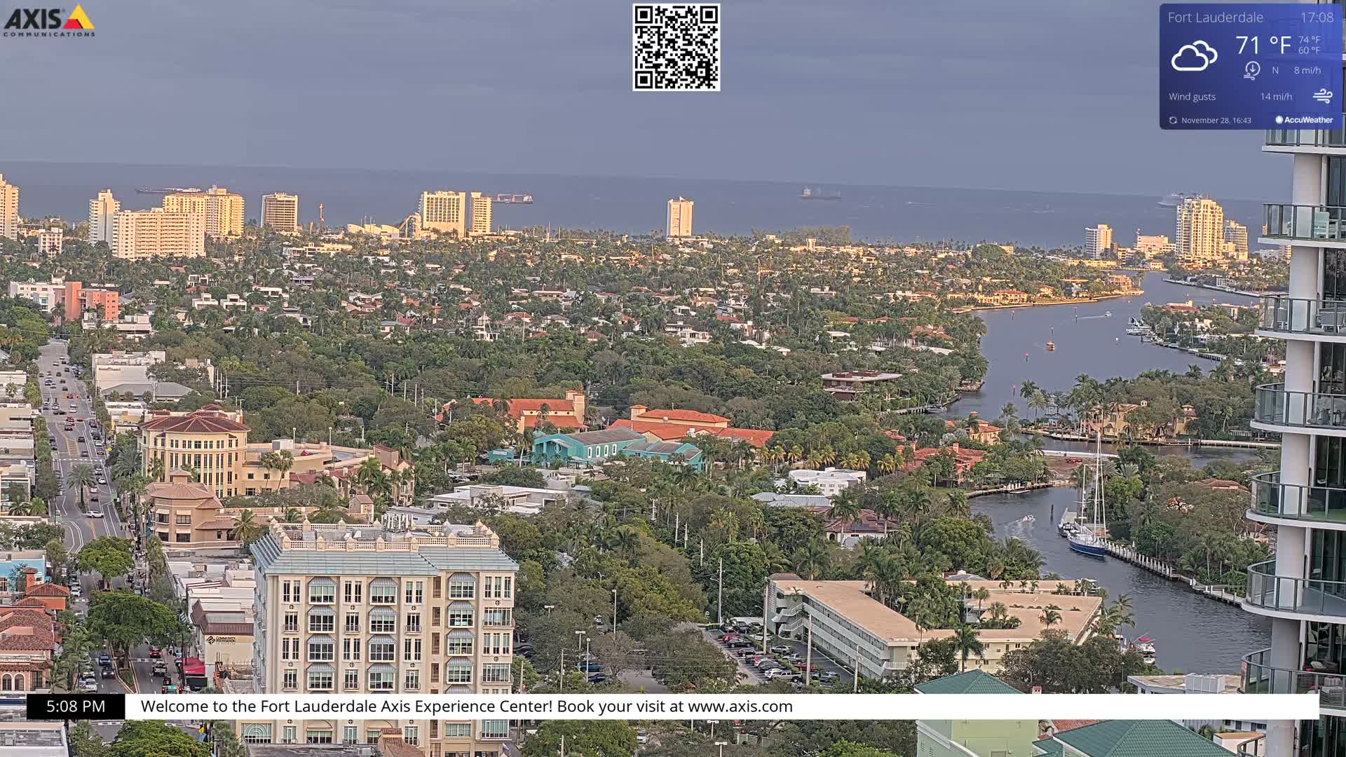 An expansive aerial view reveals a dense, tree-filled cityscape stretching to the ocean, featuring a winding river with boats and numerous buildings under a cloudy sky.
