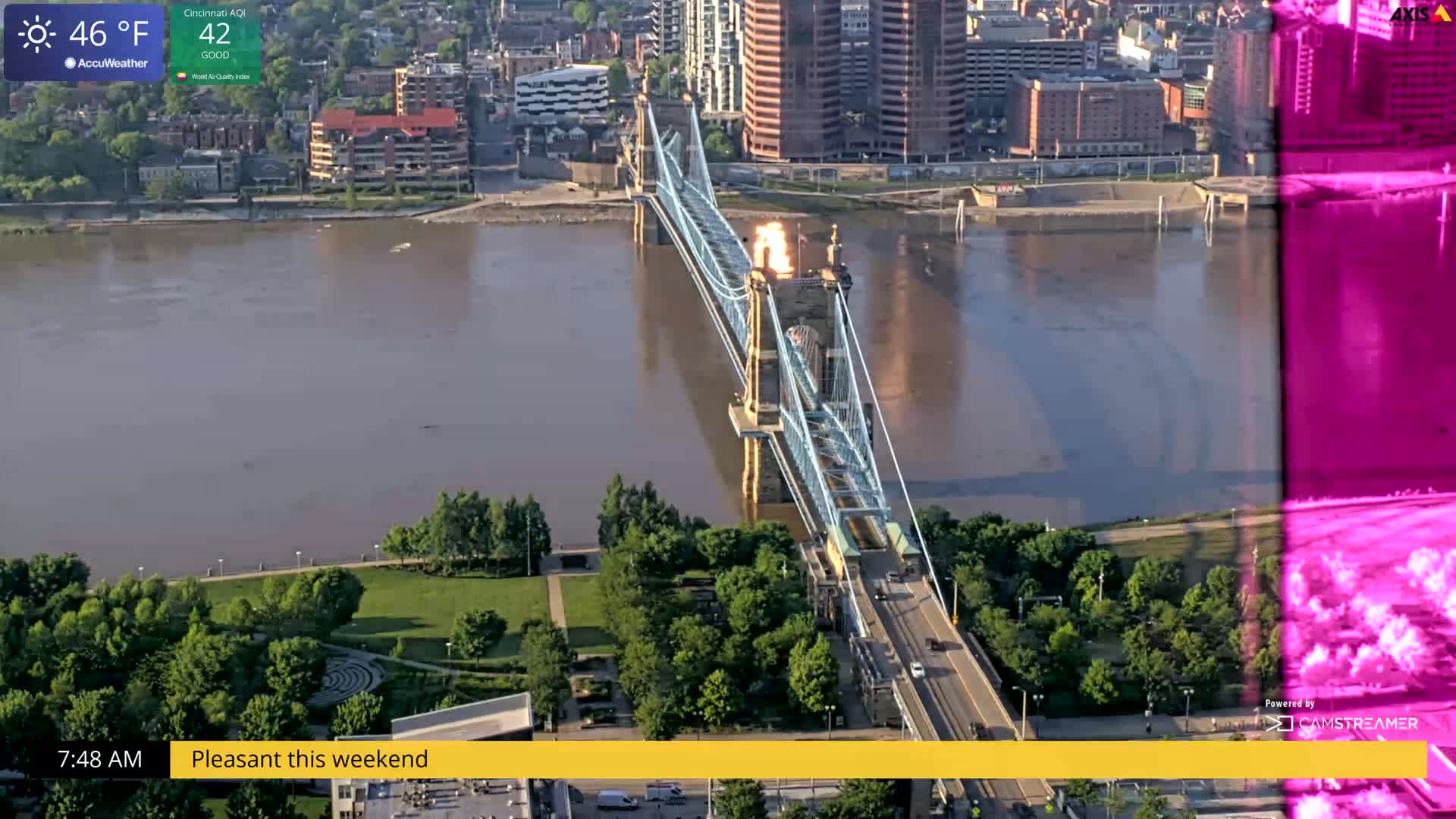 A suspension bridge spans a wide brown river, with green parkland and city buildings visible on either side, under a partly sunny sky.
