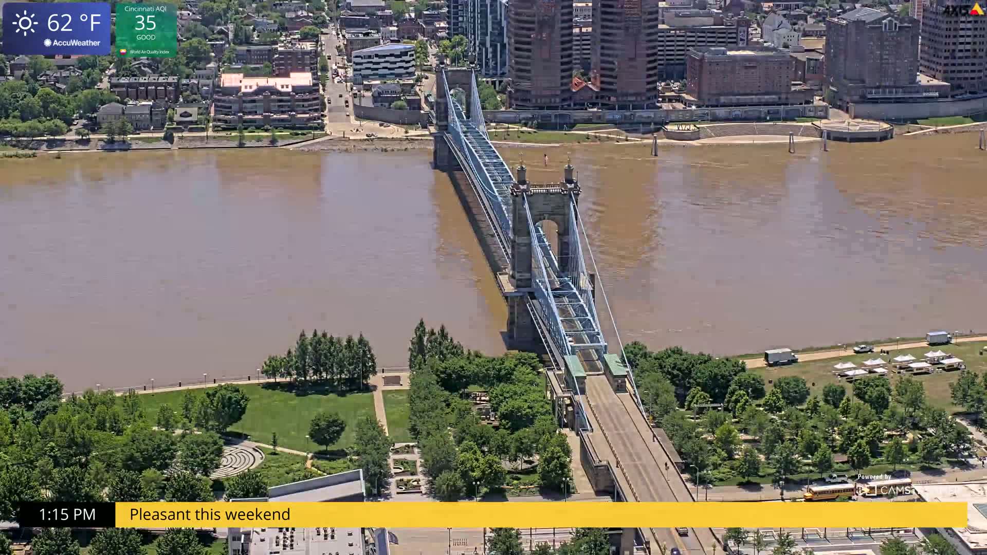 A suspension bridge spans a wide, muddy river, with green parkland and city buildings visible on either side, under sunny skies.