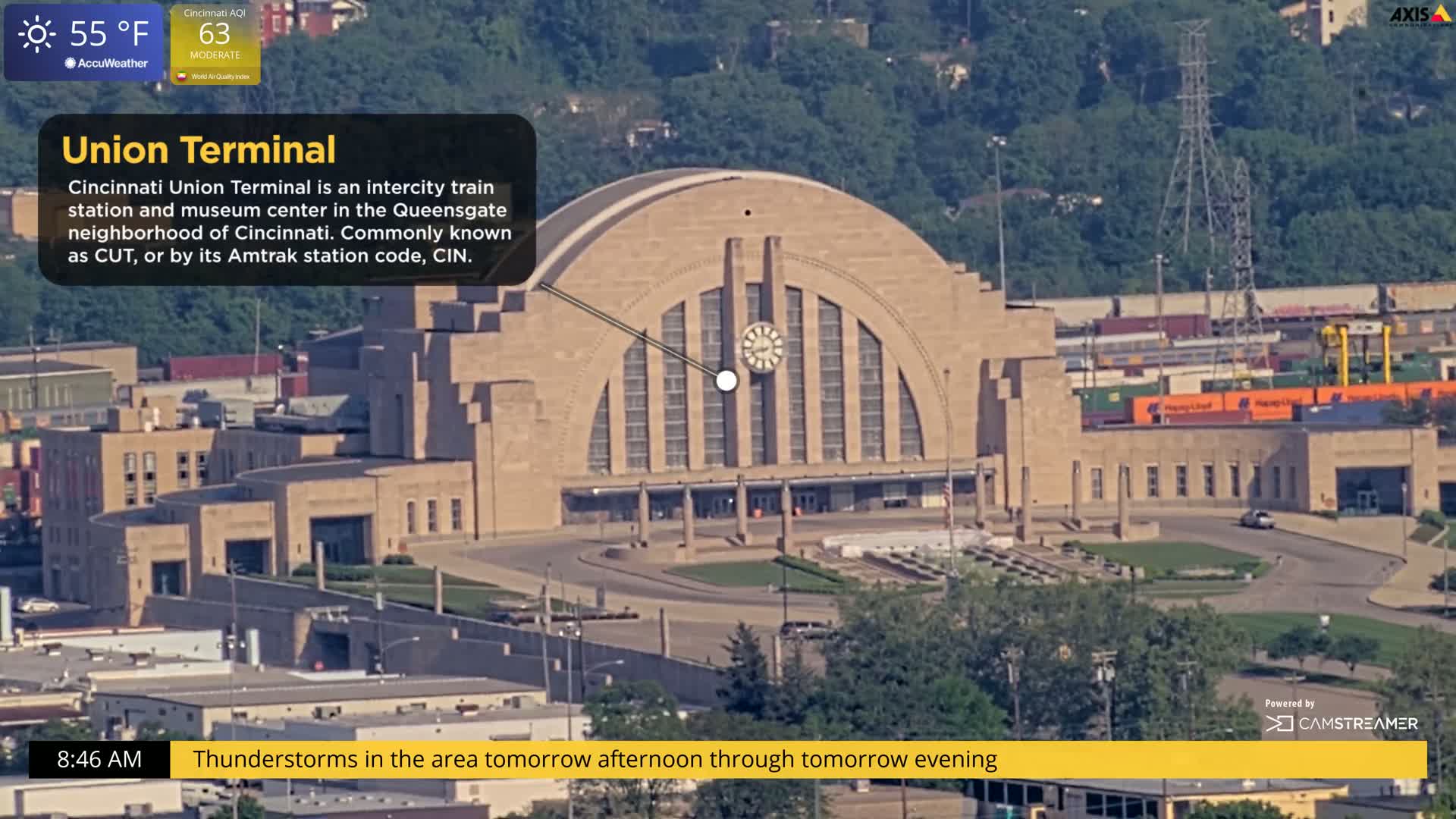 An aerial view shows a large, tan stone train station with a curved roof and clock on a partly sunny day.