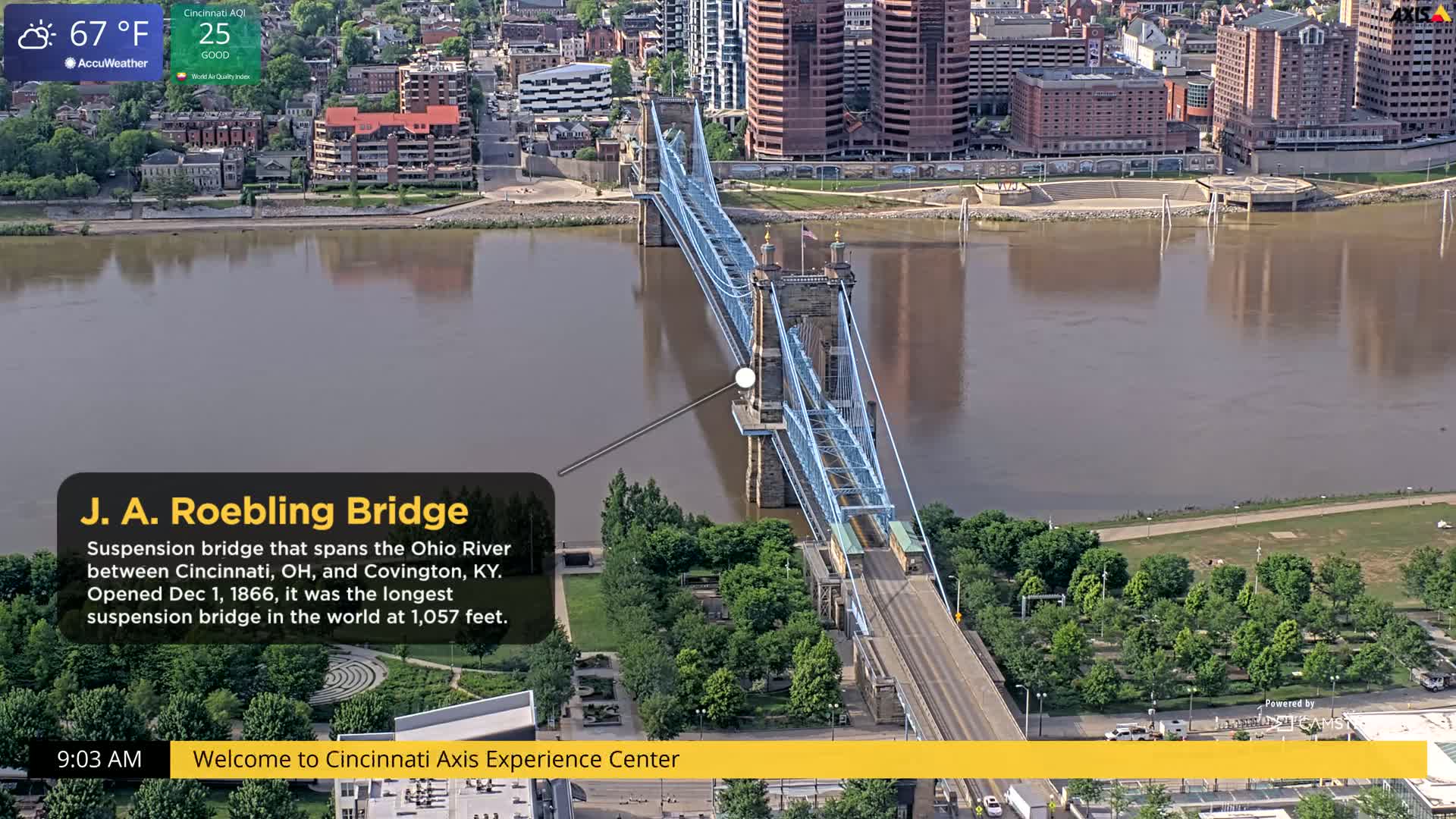A blue suspension bridge spans a wide, muddy river, with cityscapes and green spaces visible on either side under a partly cloudy sky.