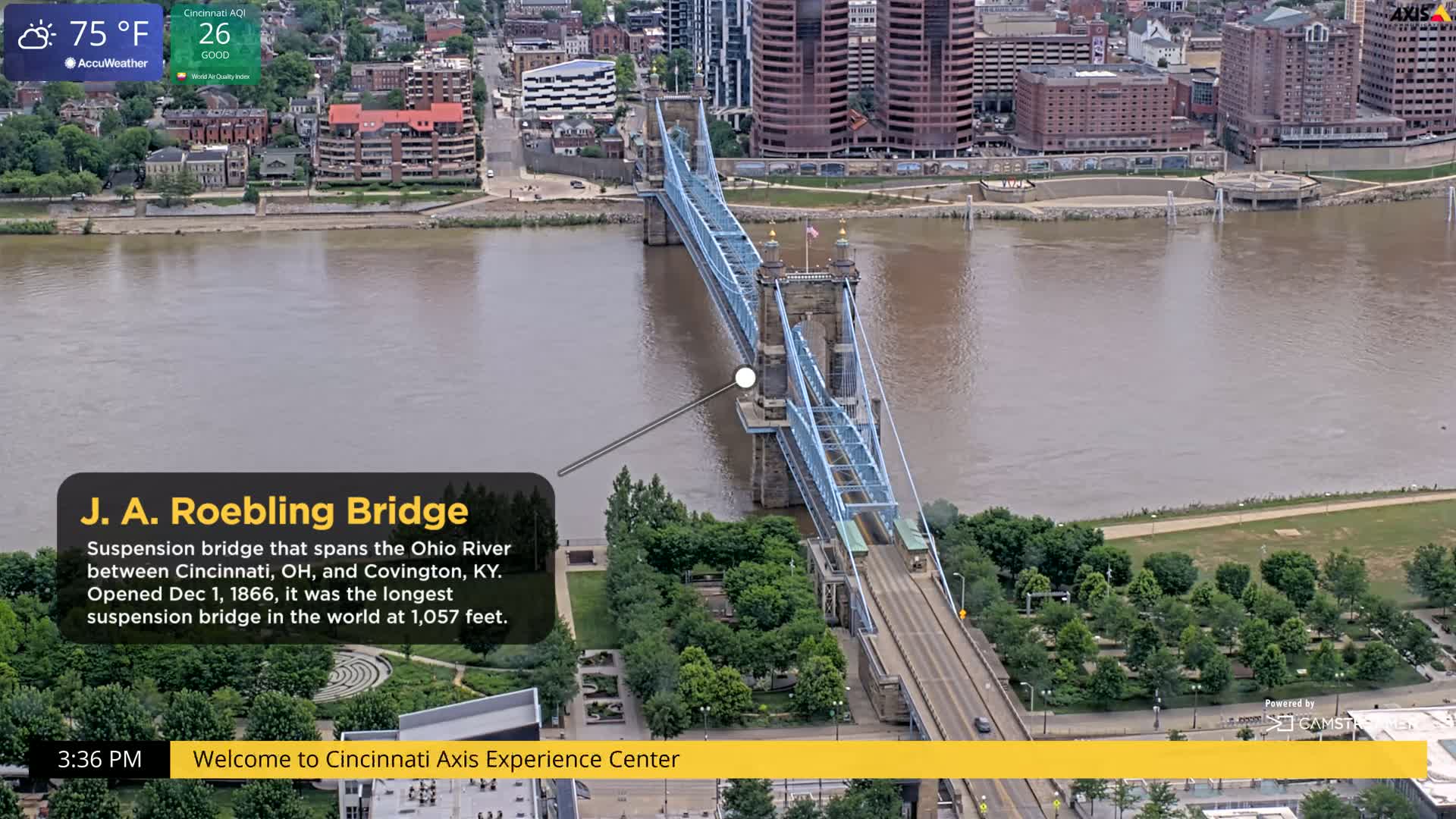 A blue suspension bridge spans a muddy brown river under a partly cloudy sky.