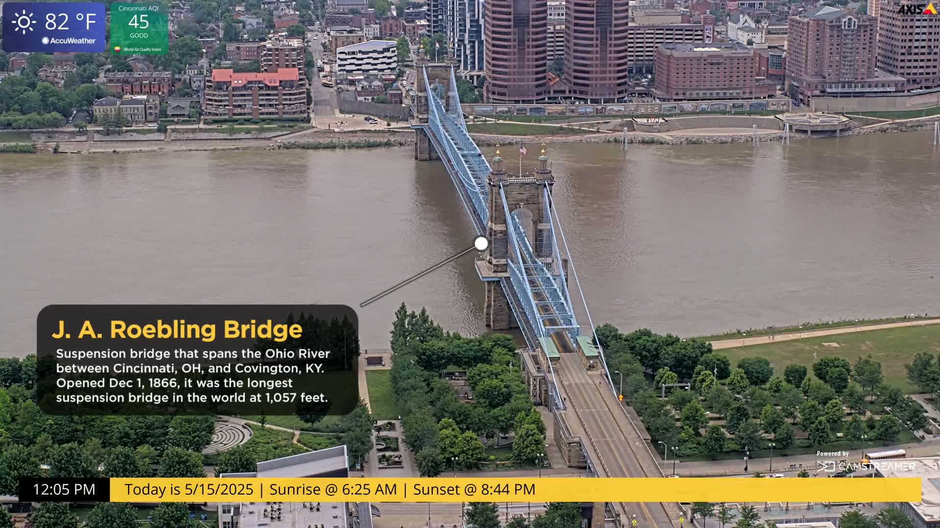 A blue suspension bridge spans a wide, muddy-brown river, surrounded by city buildings and green spaces under a partly sunny sky.