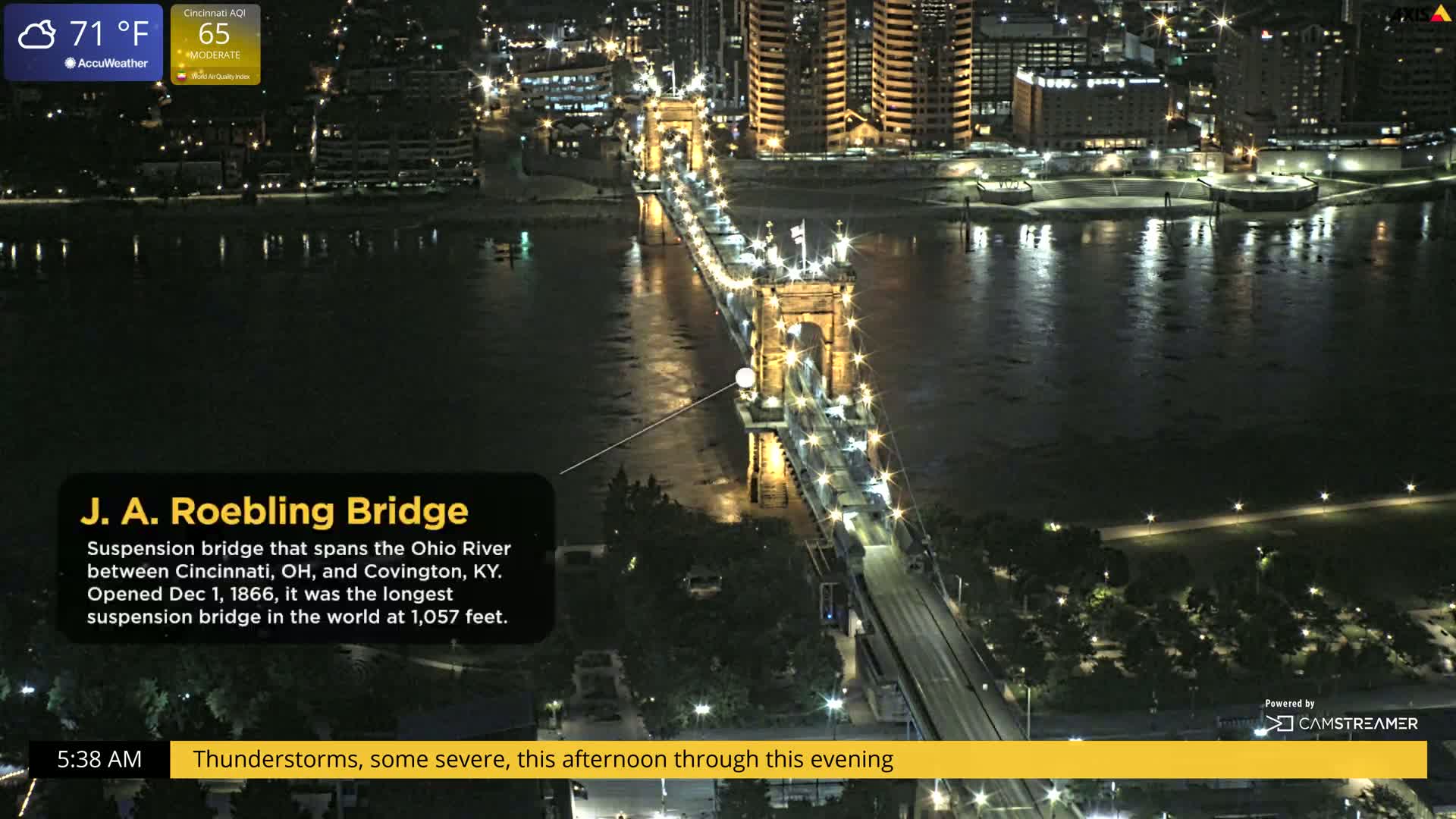 A lit suspension bridge spans a dark river at night, with thunderstorms predicted for later in the day.