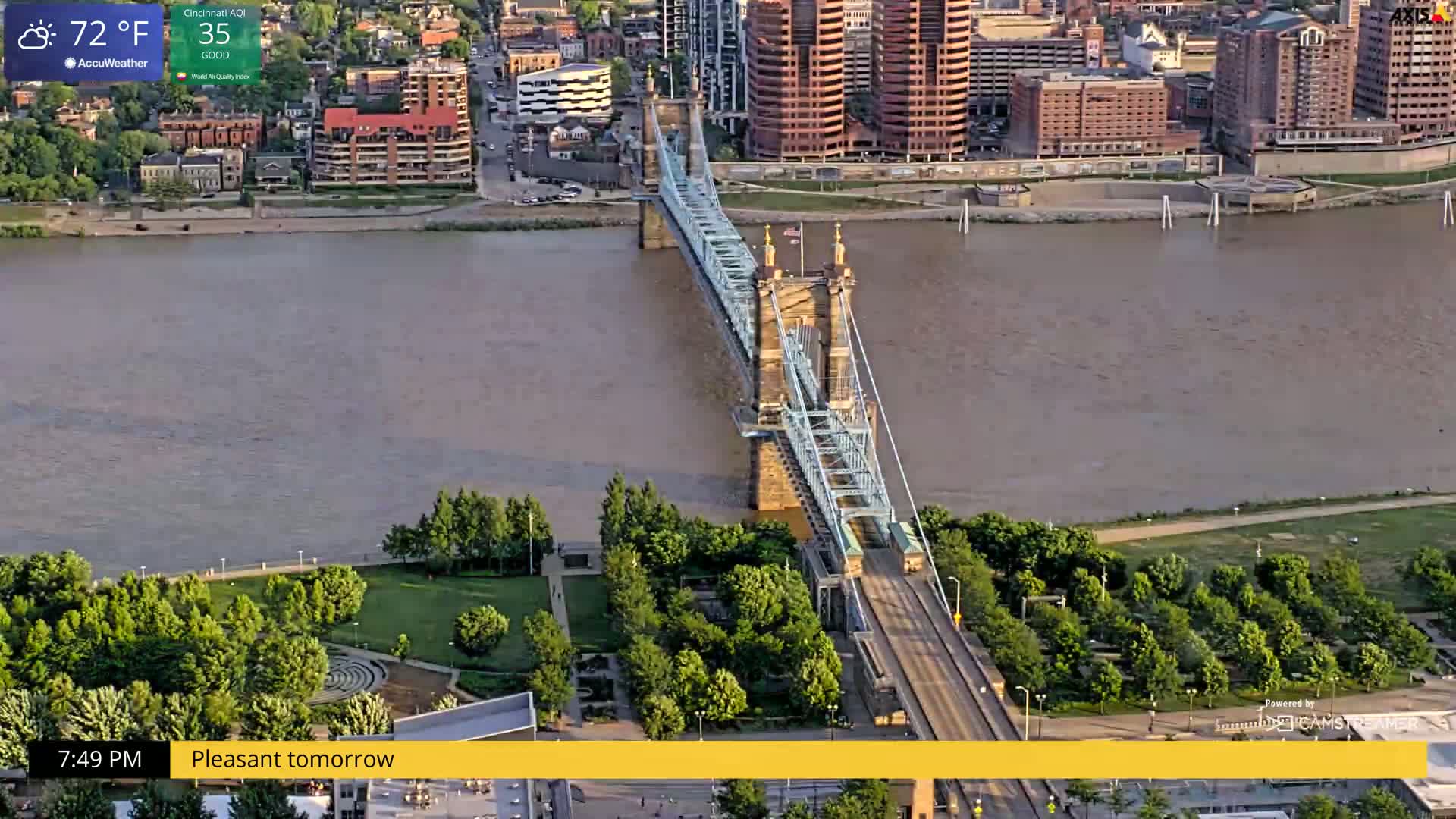 A suspension bridge spans a wide, murky river, with green parkland and city buildings visible on either side under a partly cloudy sky.