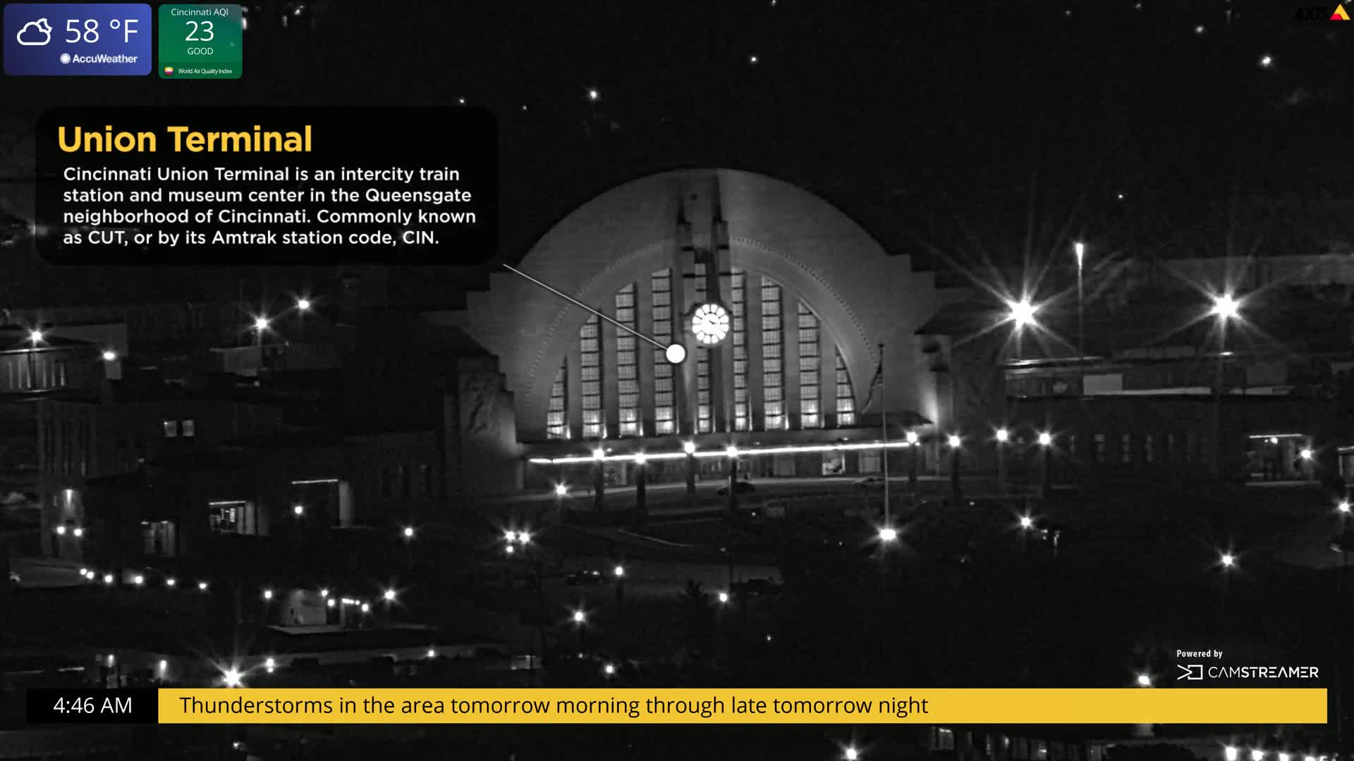 A nighttime, black-and-white view of a large, illuminated train station with a semi-circular roof, under a clear night sky, with a forecast of thunderstorms.
