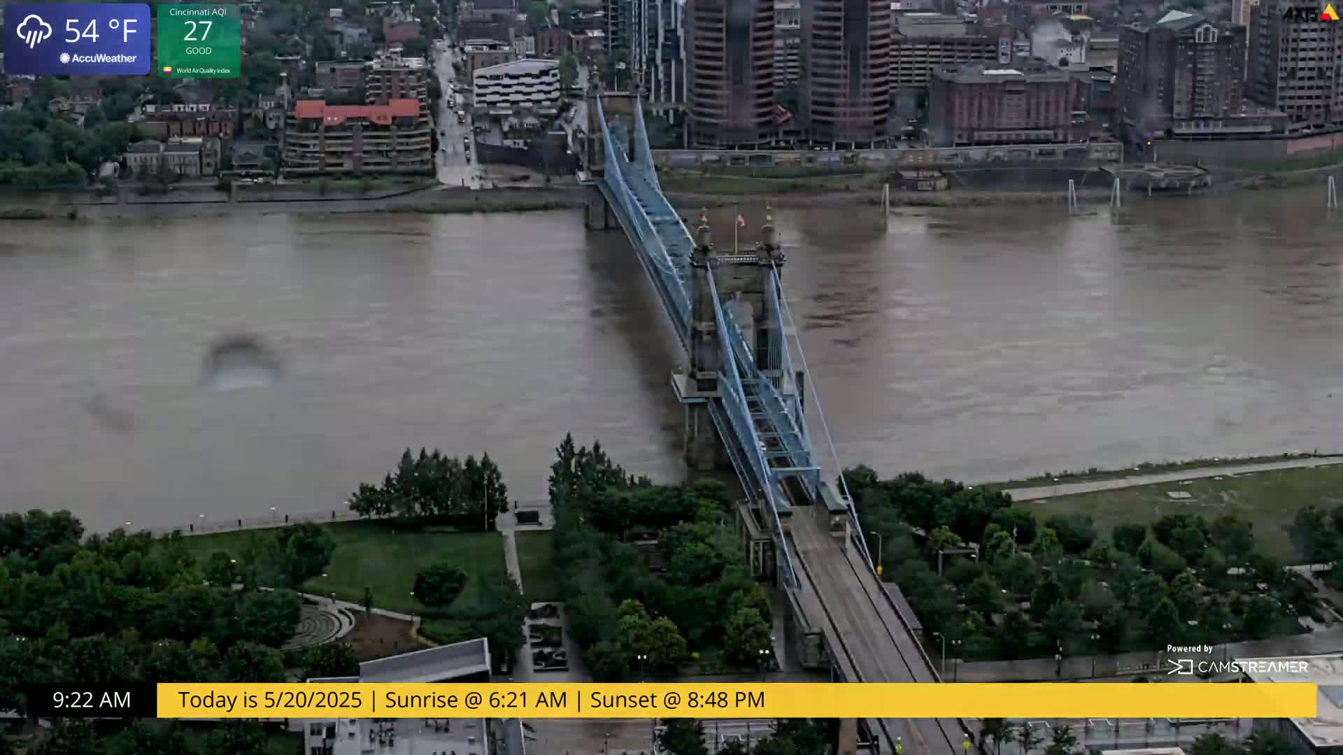 A blue suspension bridge spans a wide, muddy river under an overcast sky.