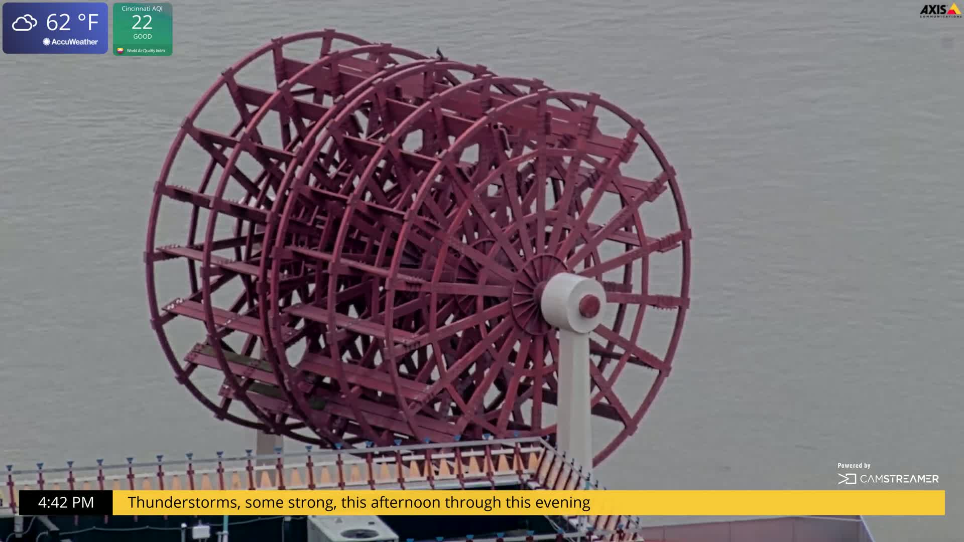 A large, reddish-brown paddle wheel sits beside a body of water under a cloudy sky with thunderstorms predicted.