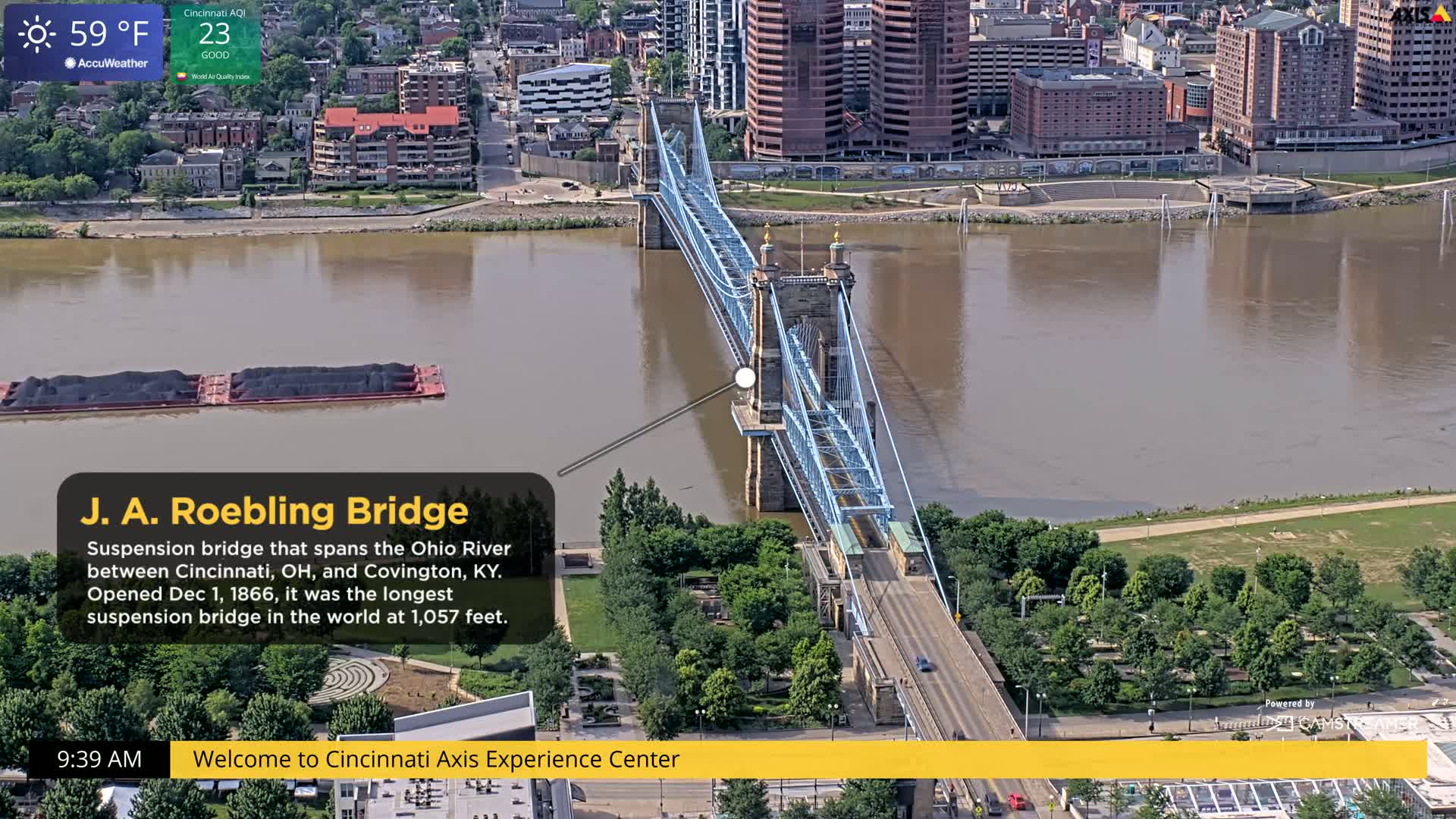 A blue suspension bridge spans a muddy-brown river, with barges on the water and green trees lining the riverbanks on a 59°F day.