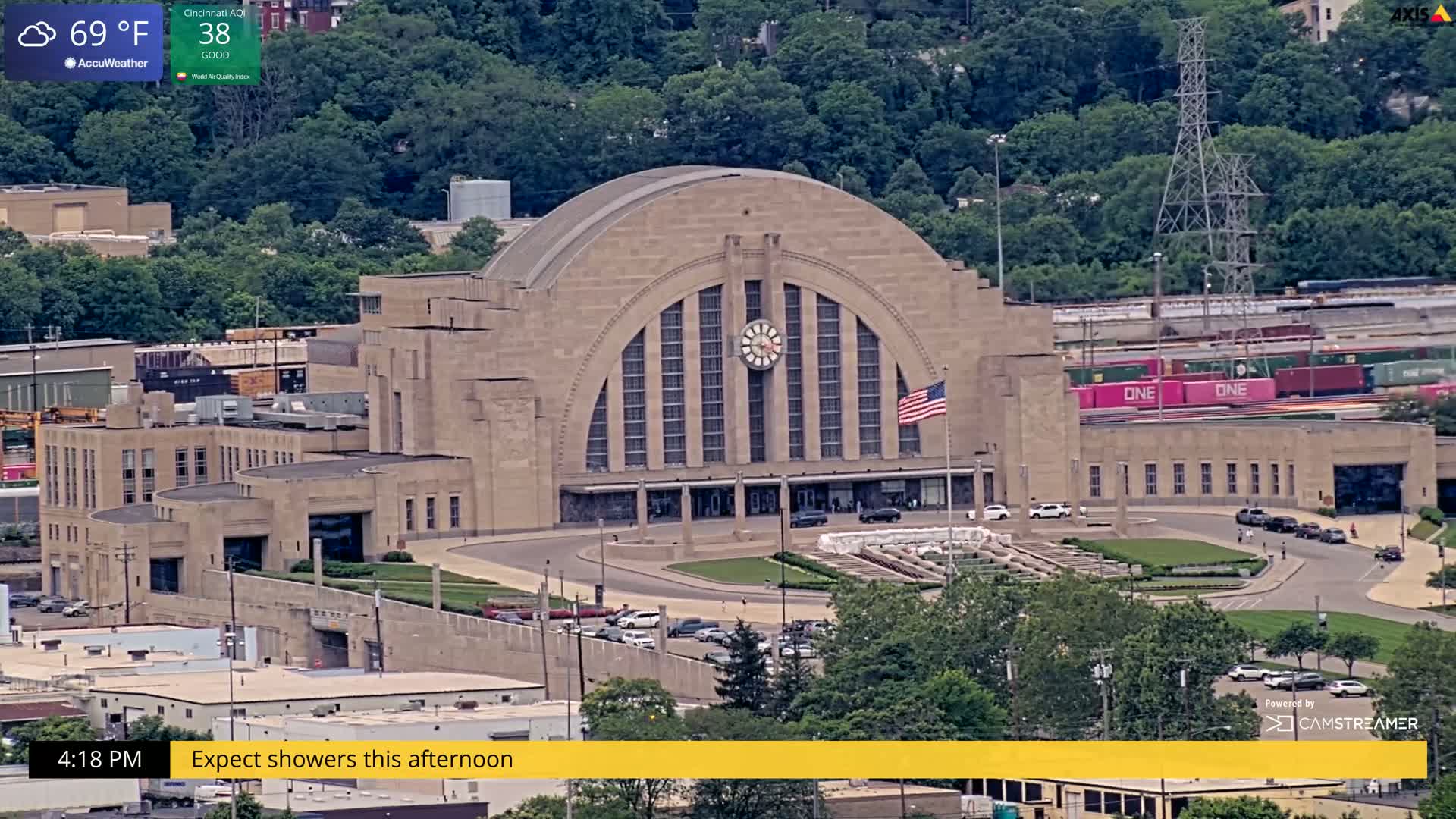 A large, tan stone building with a semi-circular roof and clock in the center is shown from an elevated view, with cars in the foreground and train cars behind it, under partly cloudy skies.