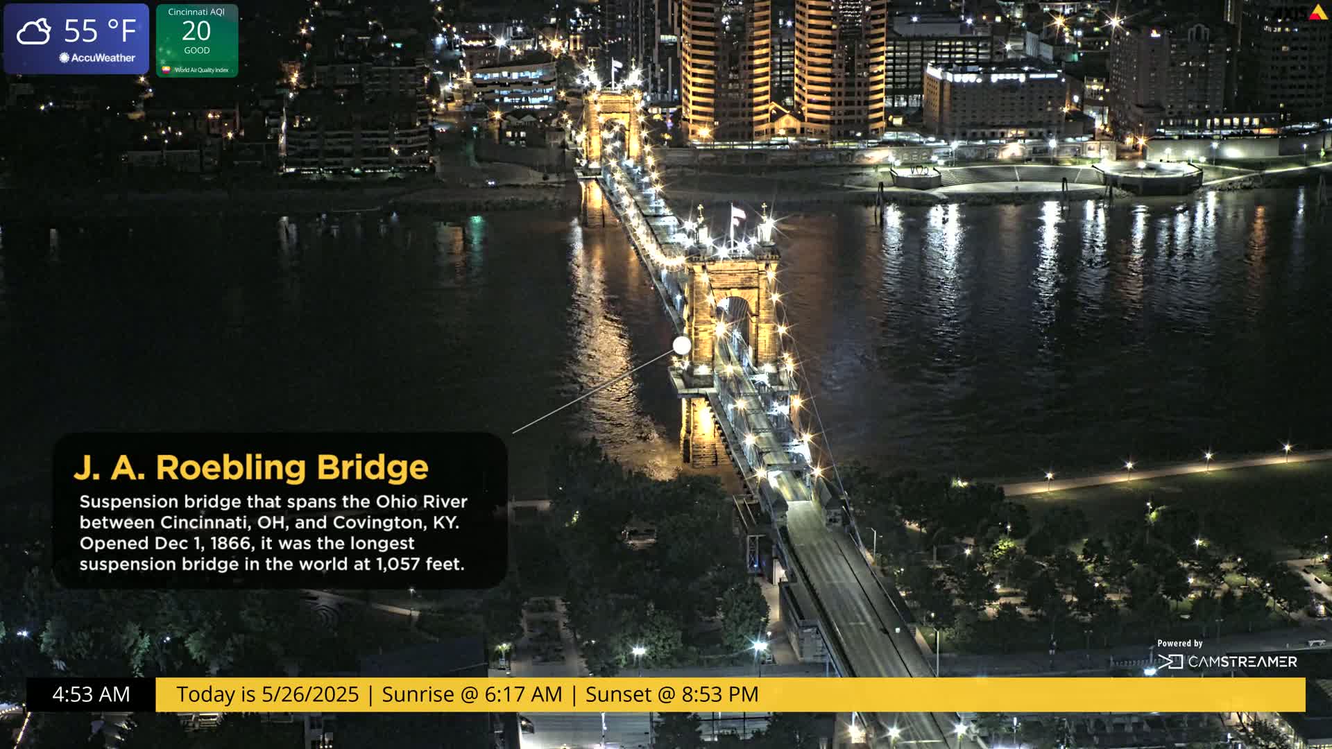 A brightly lit suspension bridge spans a dark river at night under a partly cloudy sky.