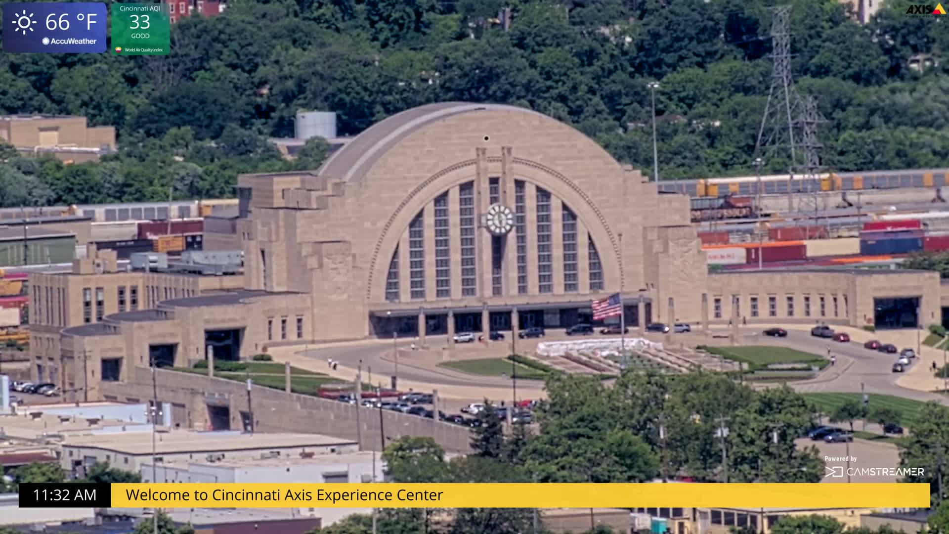 A large, tan stone building with a semi-circular front and a clock in the center is seen from an aerial view on a sunny day with a temperature of 66°F.