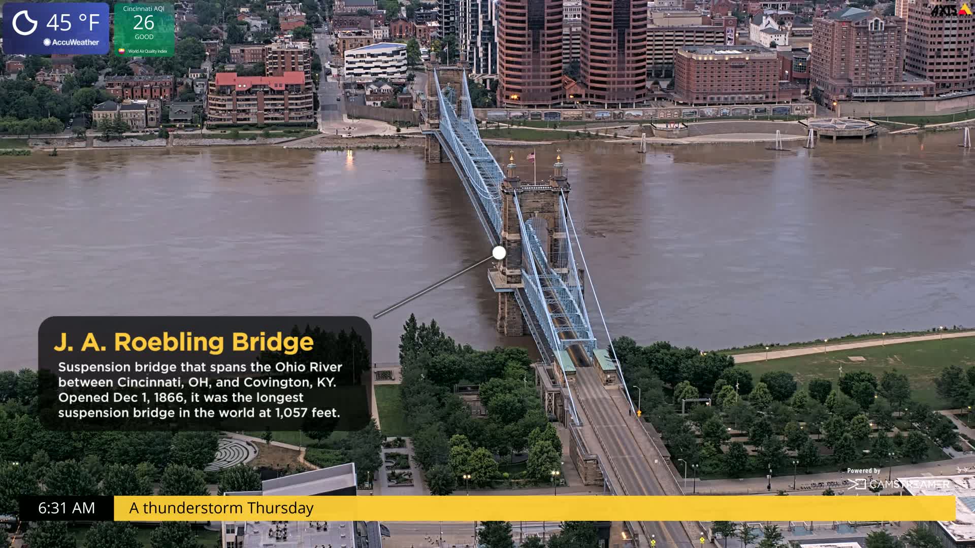 A blue suspension bridge spans a muddy brown river under a cloudy sky during a thunderstorm.