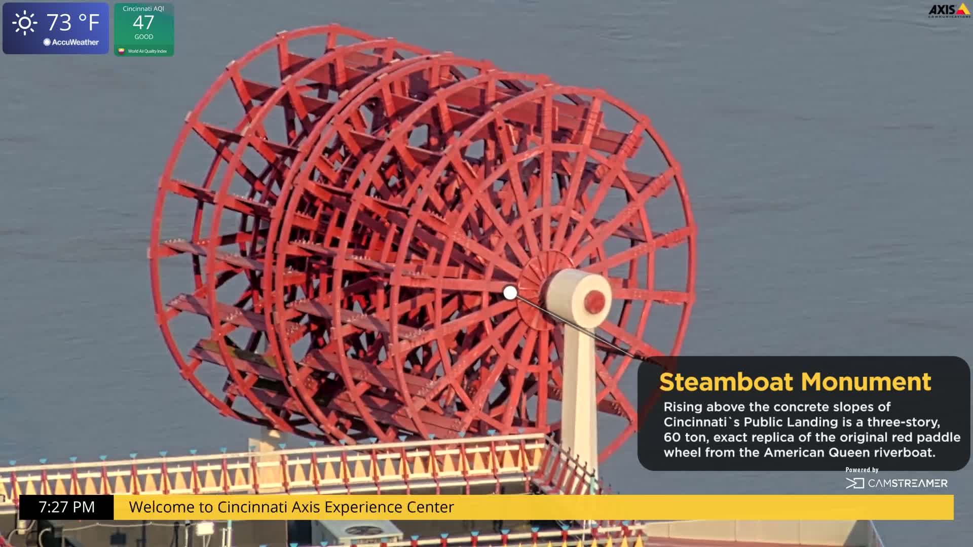 A large, red paddle wheel rises above a concrete structure next to a body of calm, gray water.