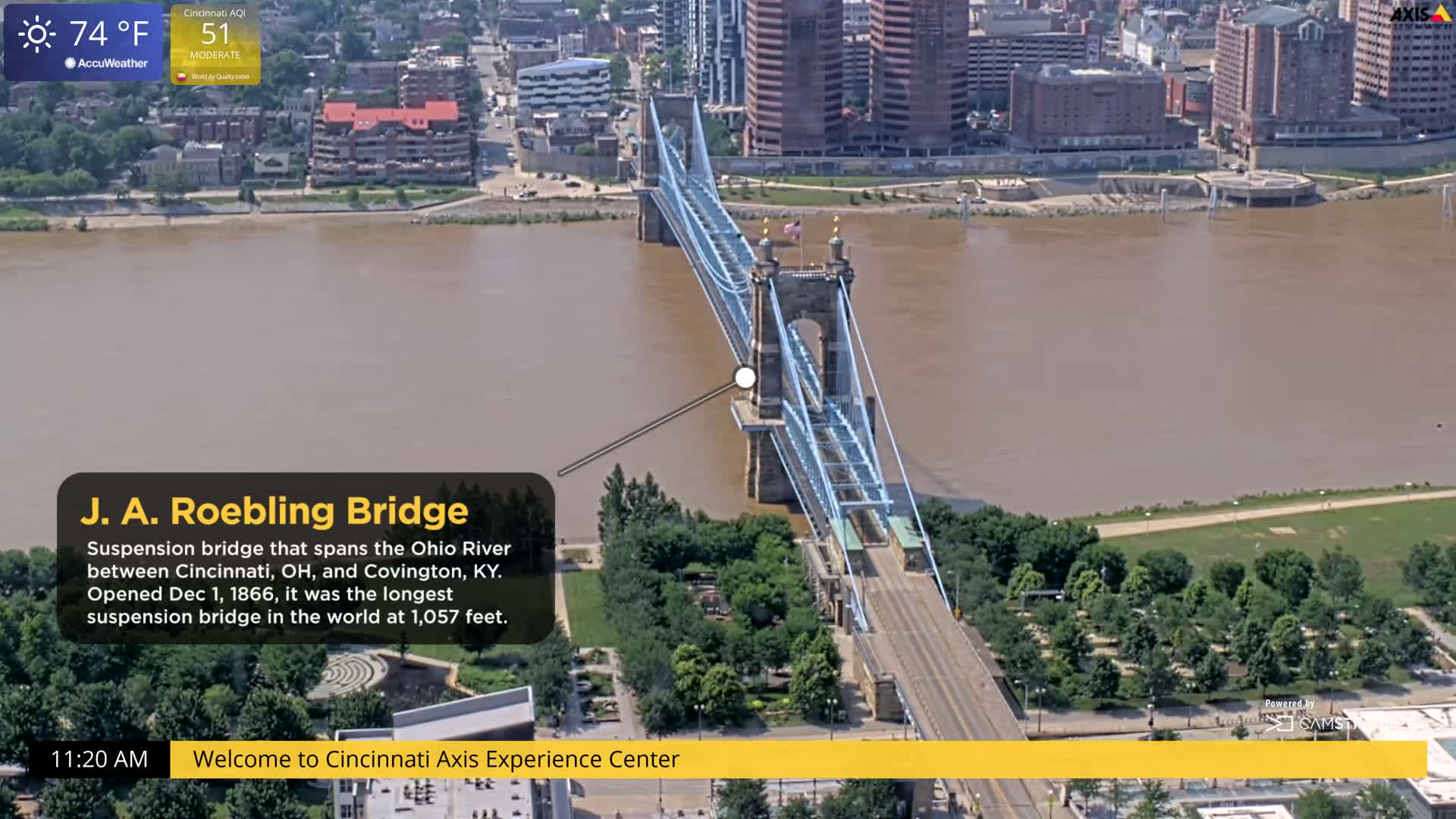 An aerial view shows the J. A. Roebling Suspension Bridge spanning a muddy-brown river, on a sunny day with a temperature of 74°F.
