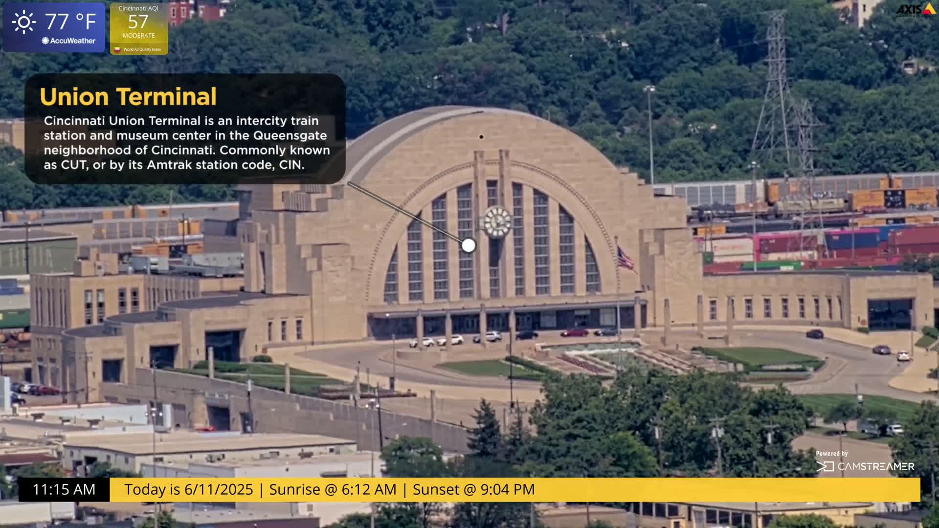 An aerial view shows a large, tan-colored train station on a partly sunny day.