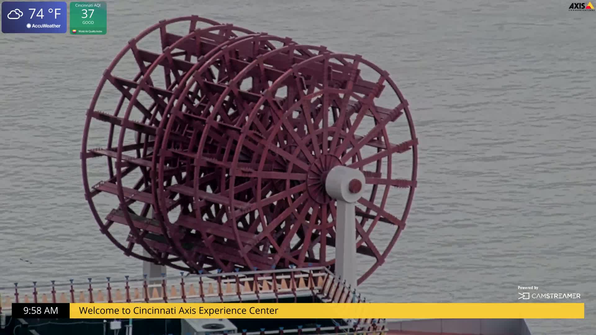 A large, reddish-brown paddle wheel sits by a body of calm, gray water on a partly cloudy 74°F day.