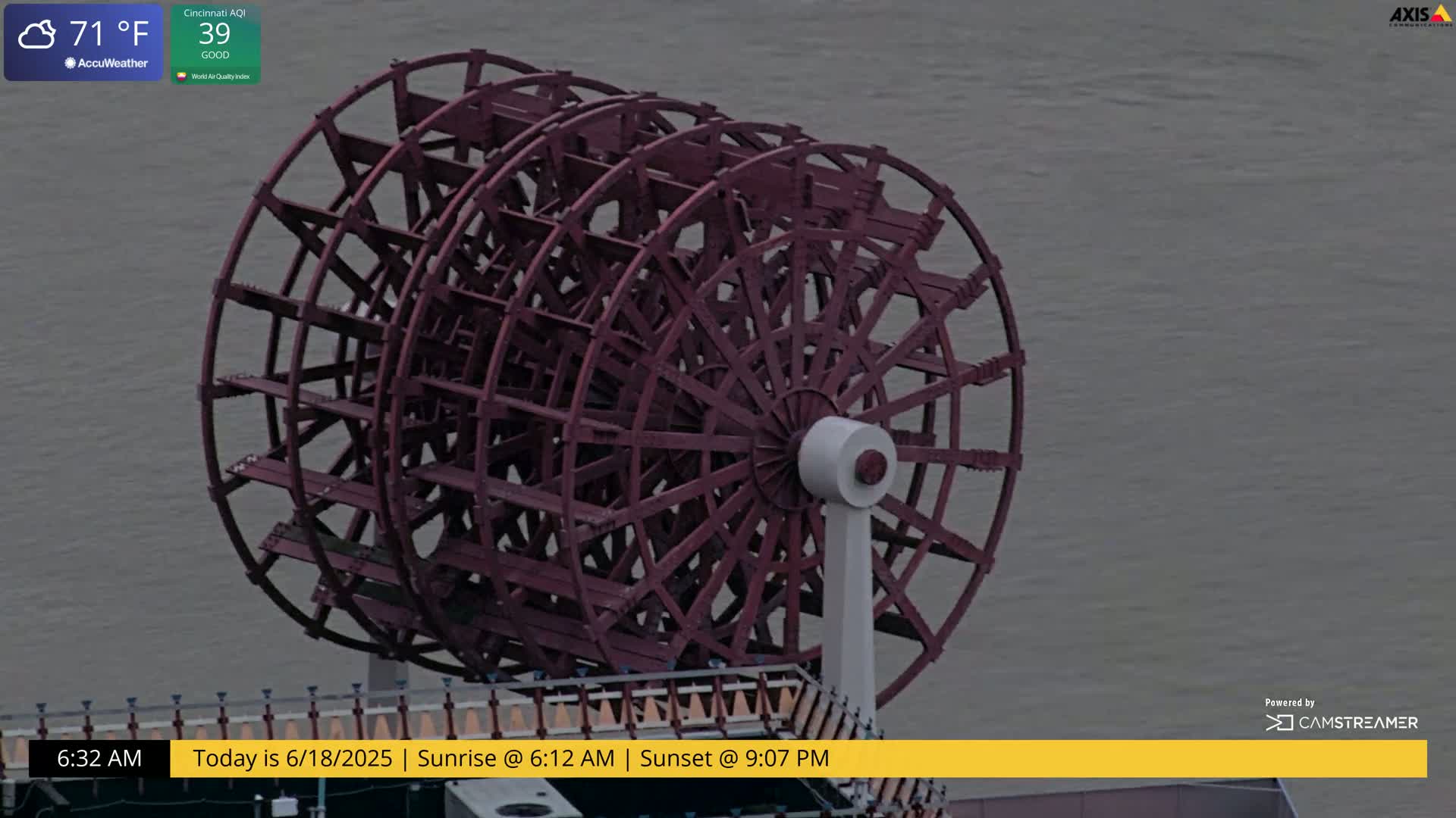 A large, dark-red paddle wheel sits beside a body of calm, gray water under cloudy skies.