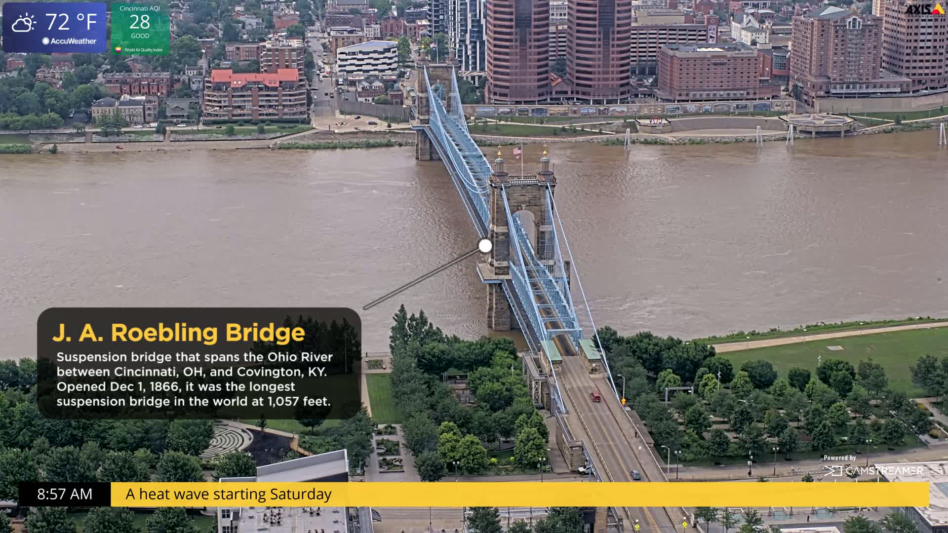 A blue suspension bridge spans a wide, muddy river under a partly cloudy sky.
