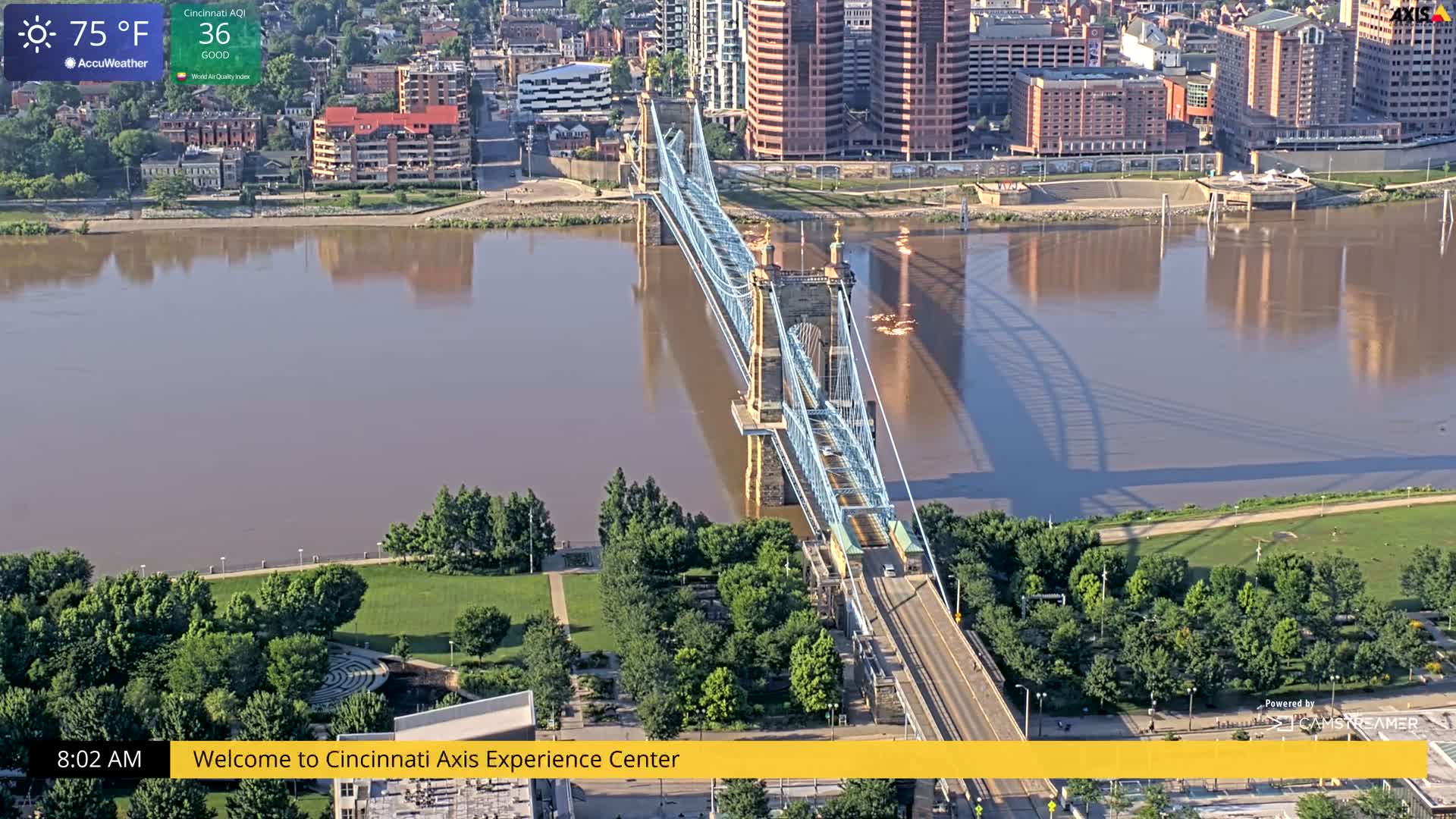 A suspension bridge spans a wide, muddy river under a sunny sky, with green parkland and city buildings visible on either side.