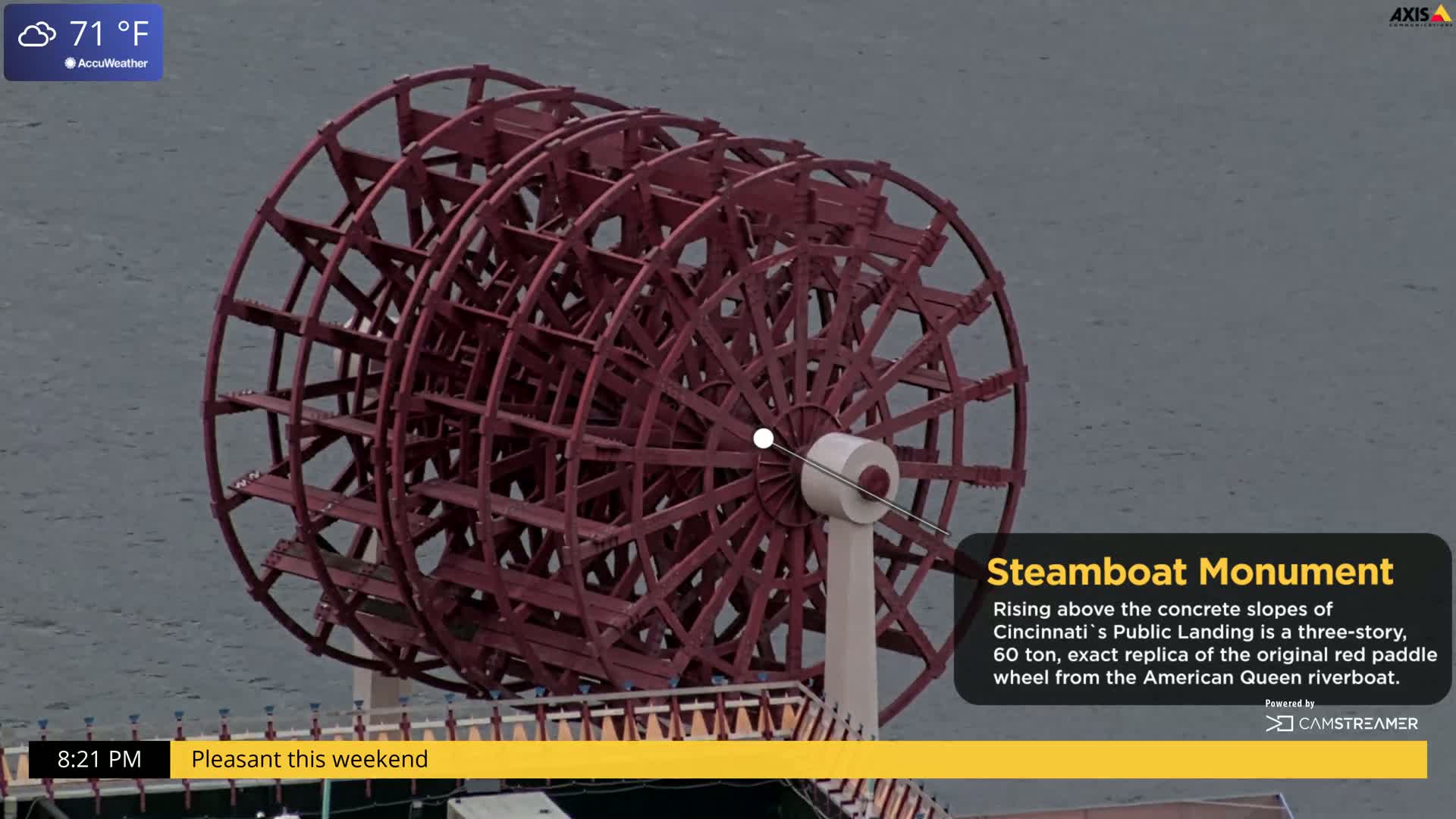 A large, reddish-brown steamboat paddle wheel rises above a body of water under partly cloudy skies.